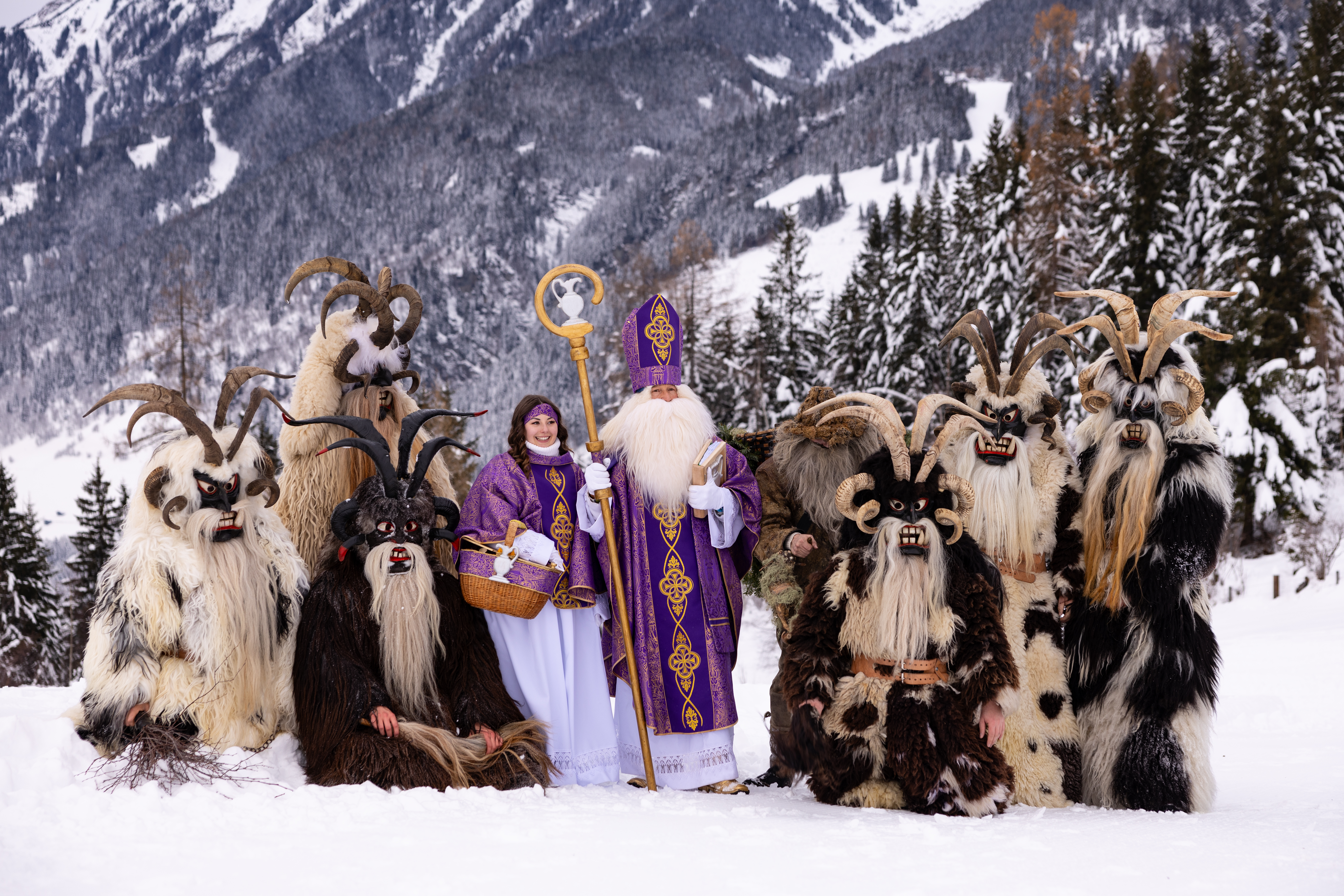 A snowy landscape featuring characters in costumes, including a robed figure and several horned, furry beings.