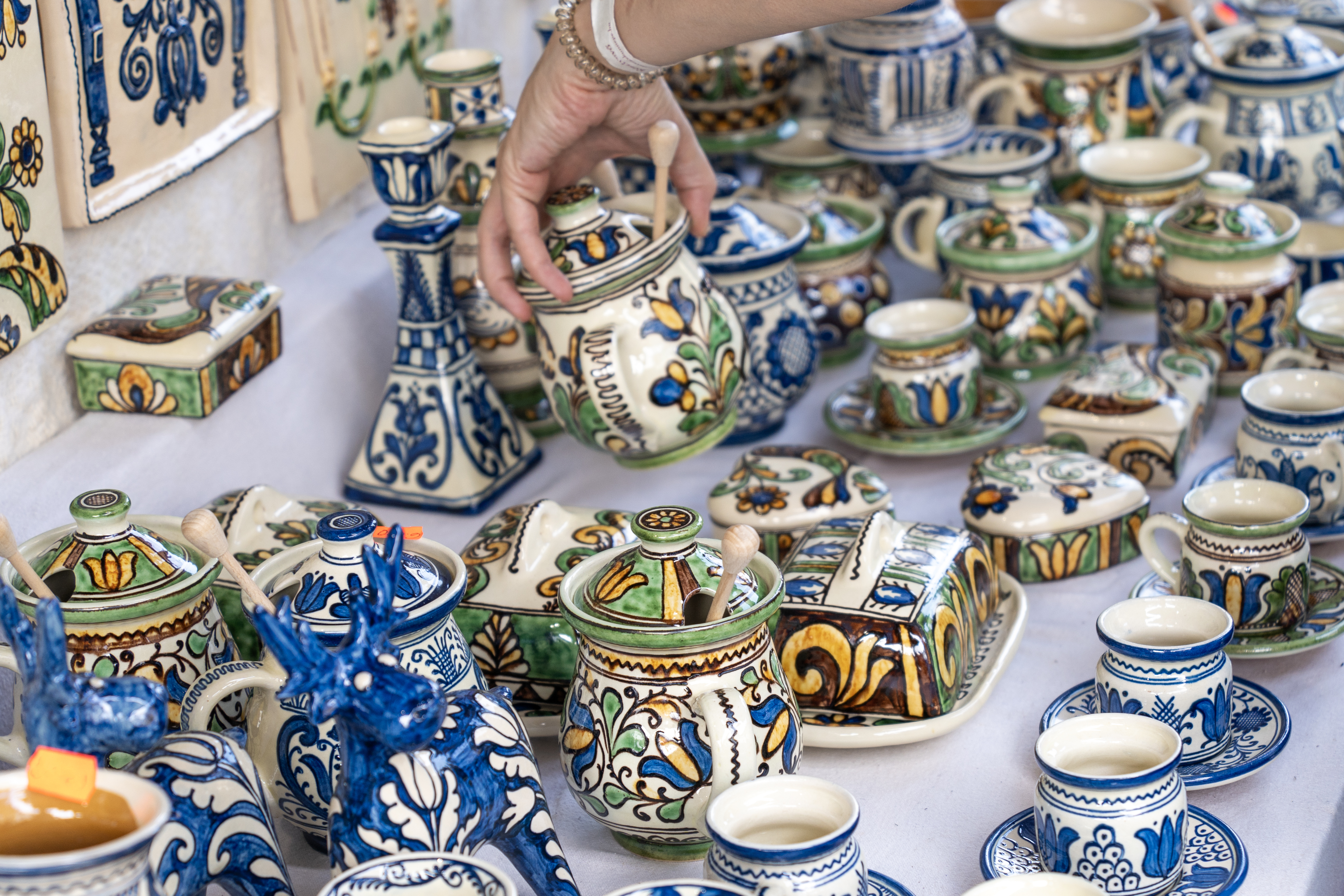A hand reaches for a colorful, ornate pottery jar among various other decorated ceramics on a display table.