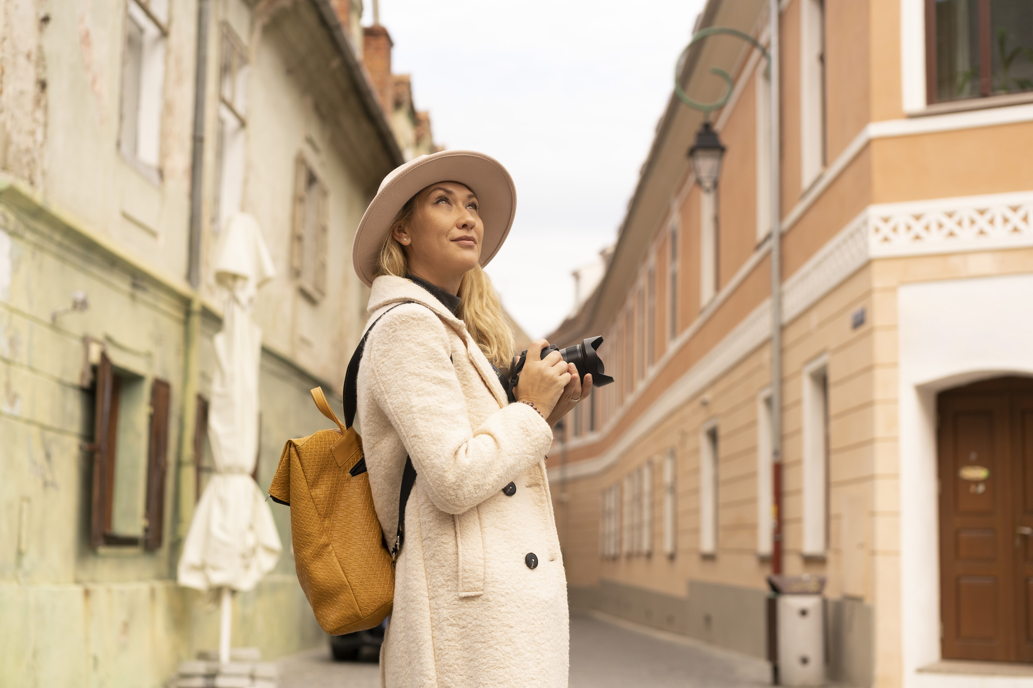 A woman with a camera looks up while standing in a charming, narrow street with colorful buildings.