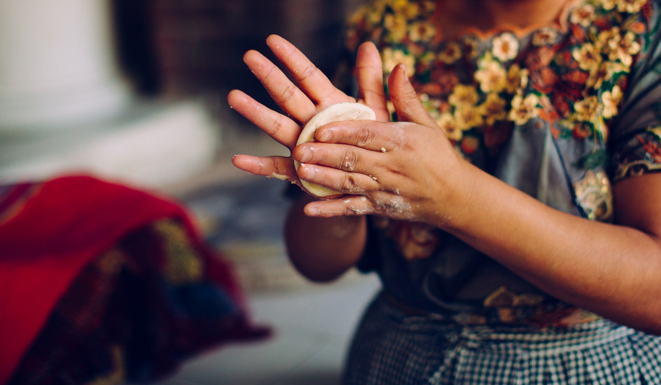 A person is shaping dough in their hands, wearing a colorful floral top with a checkered apron.
