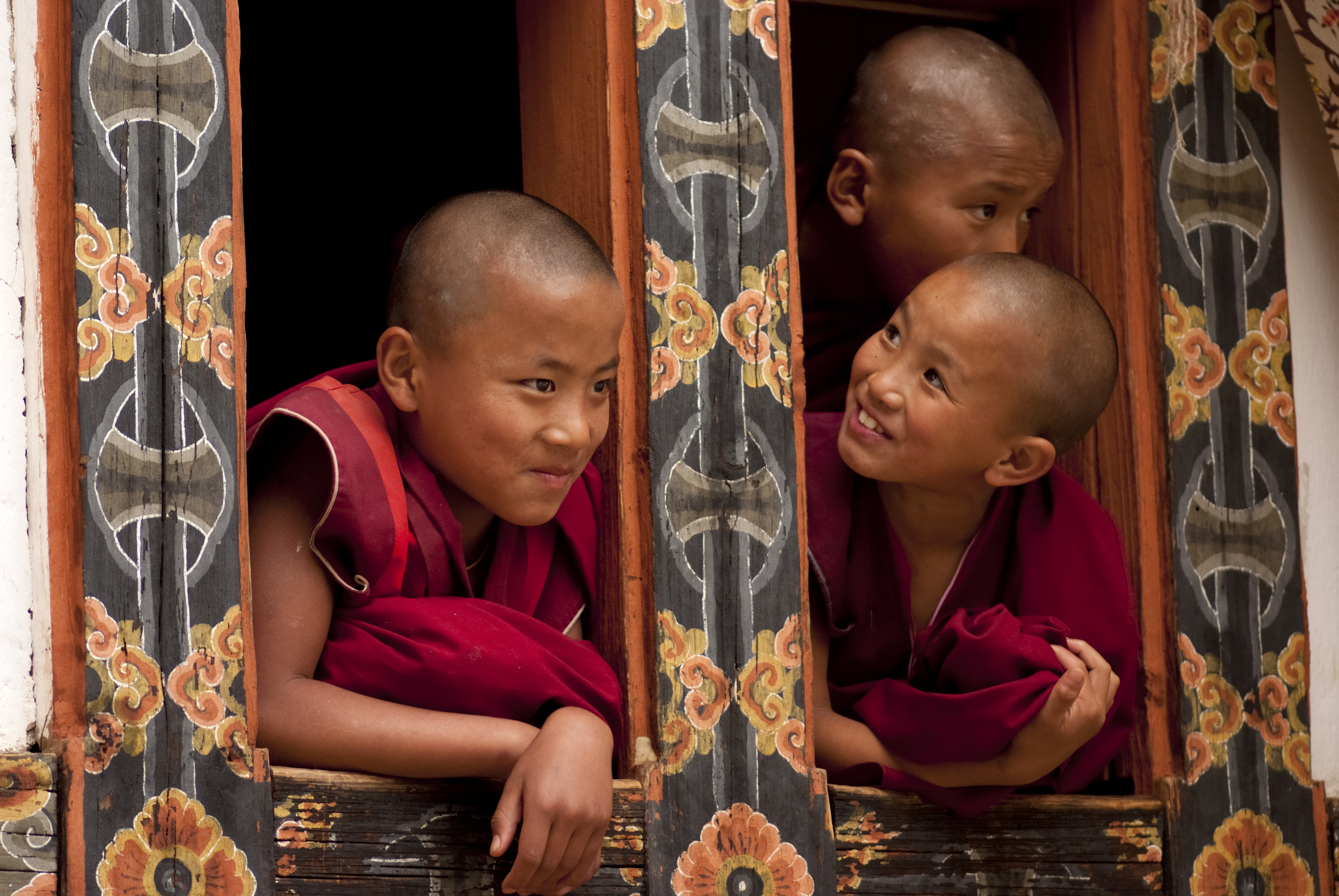 Three young monks with shaved heads peek out from a beautifully decorated window, smiling and engaging with each other.