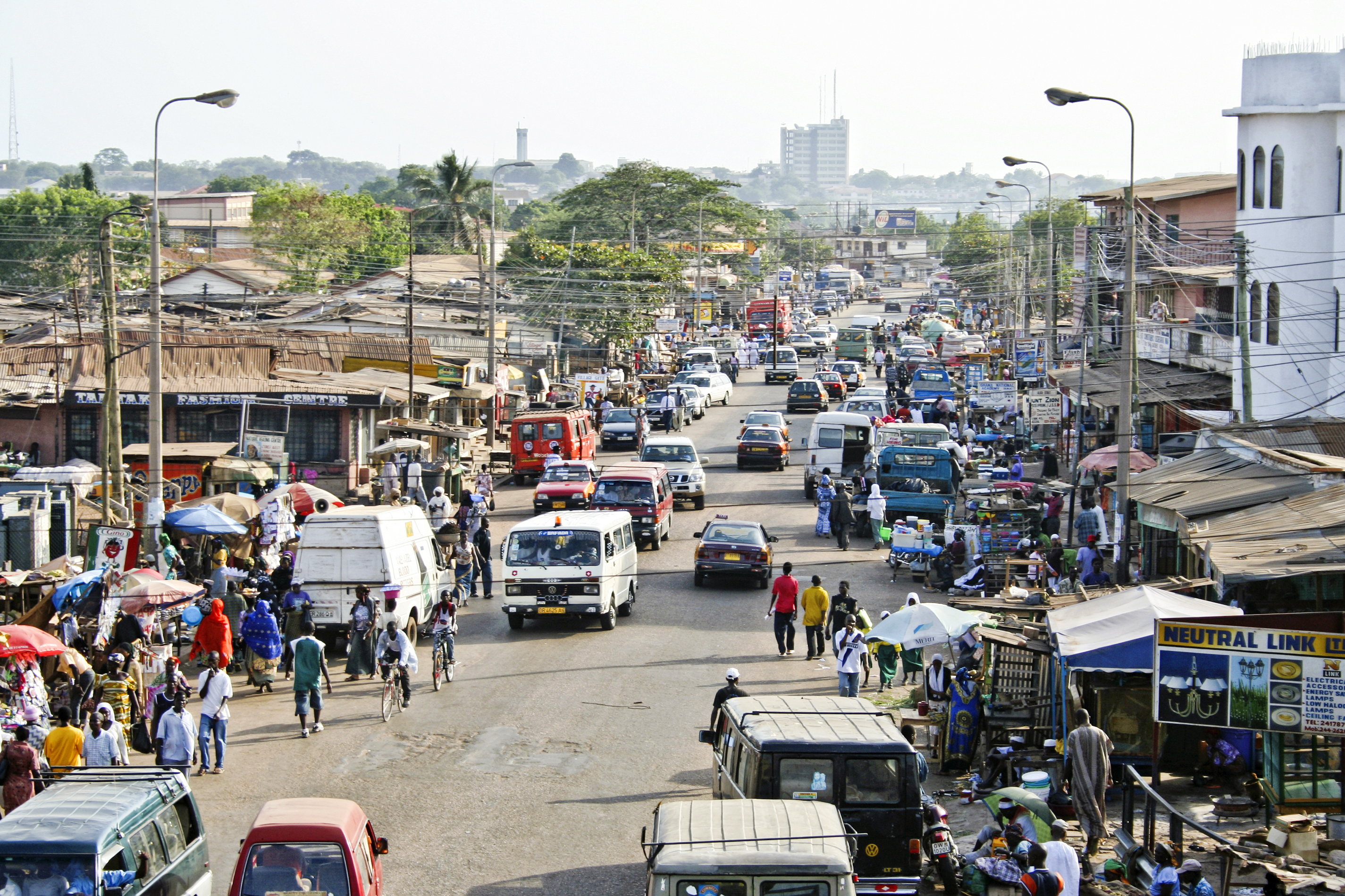 Busy street scene in a town, filled with vehicles, vendors, and pedestrians amidst shops and buildings.