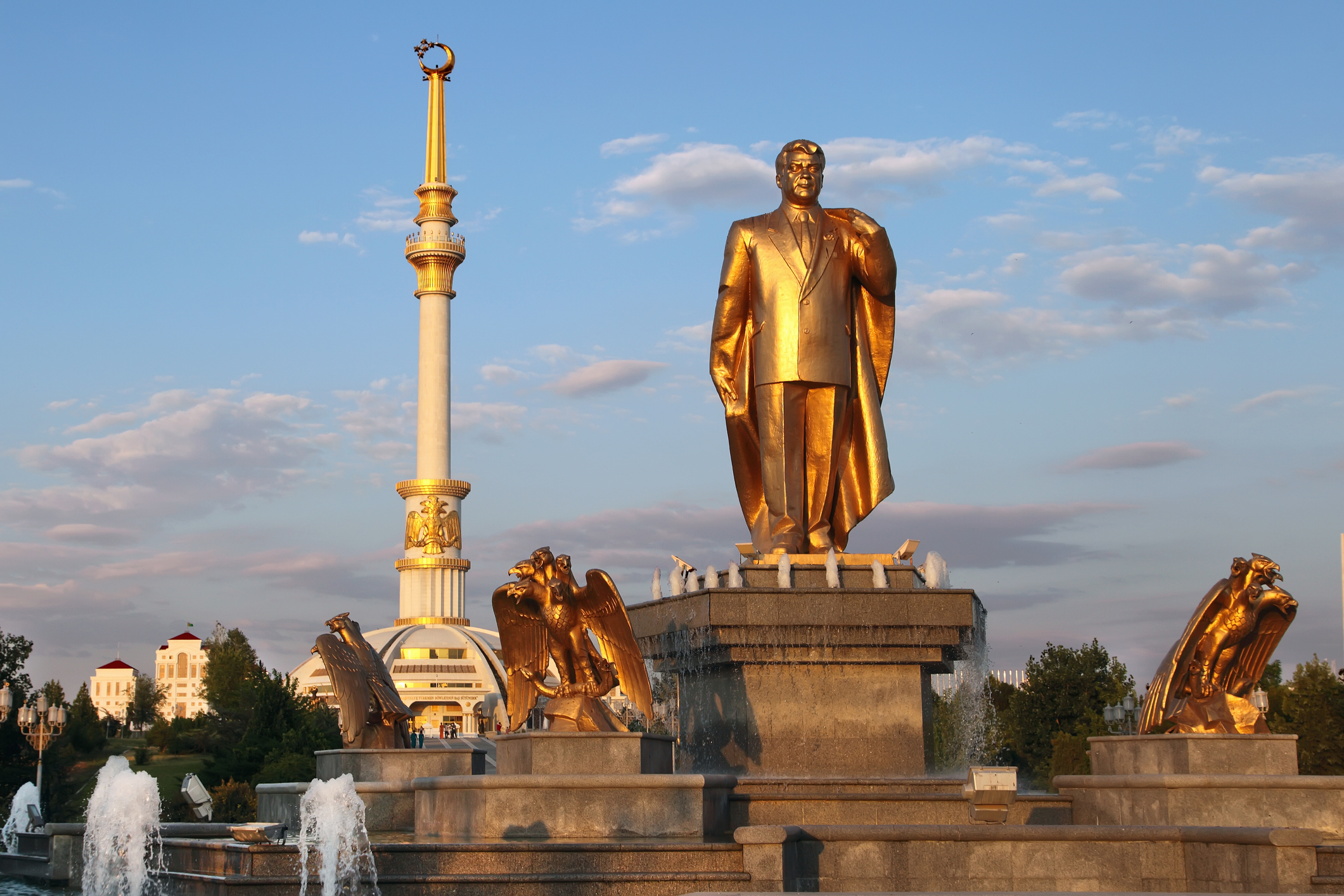 Golden statue of a man with a cape, surrounded by fountains, and a tall spire in the background against a blue sky.