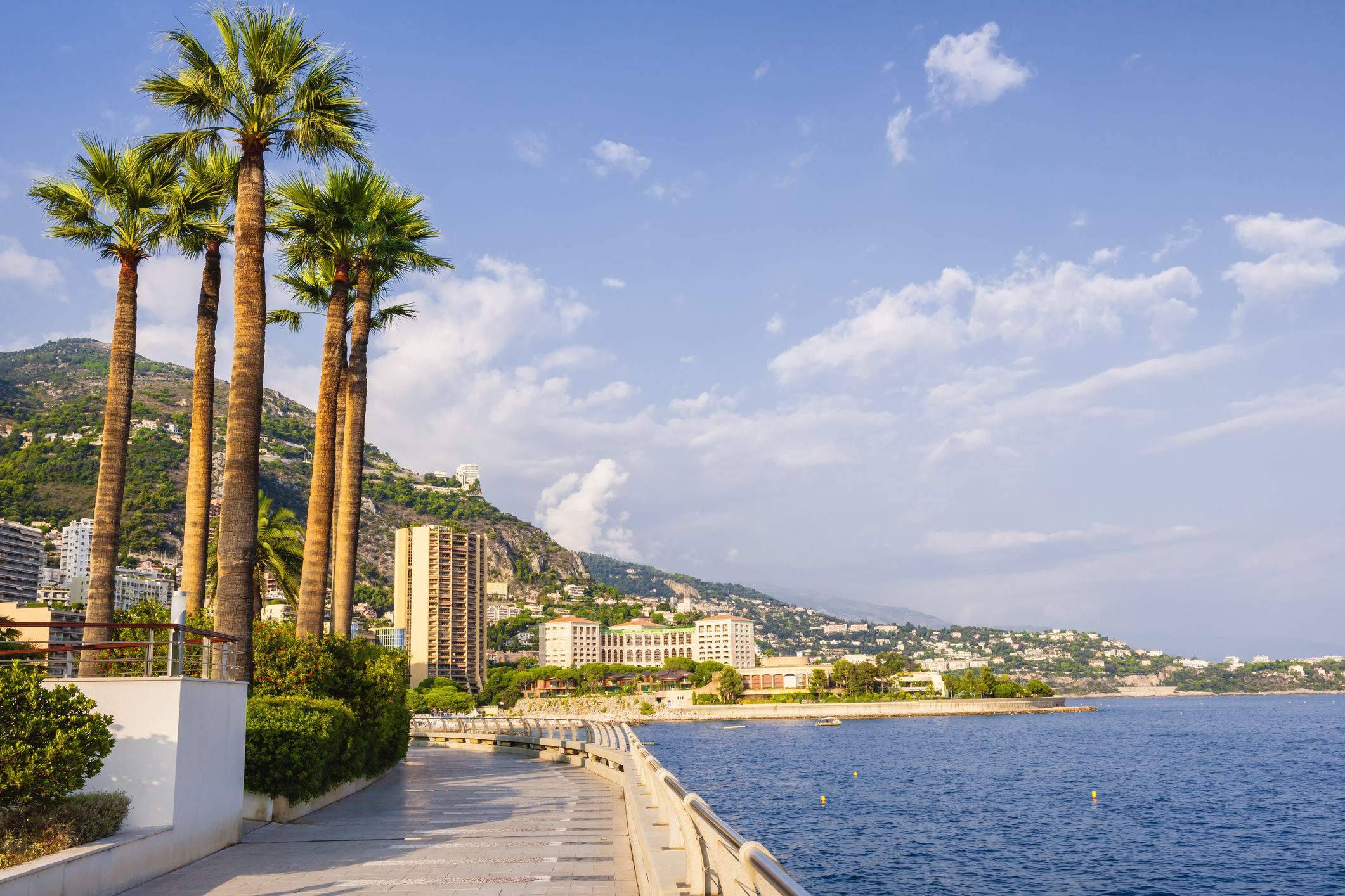 Scenic view of a coastal promenade lined with palm trees, buildings, and mountains under a blue sky with clouds.