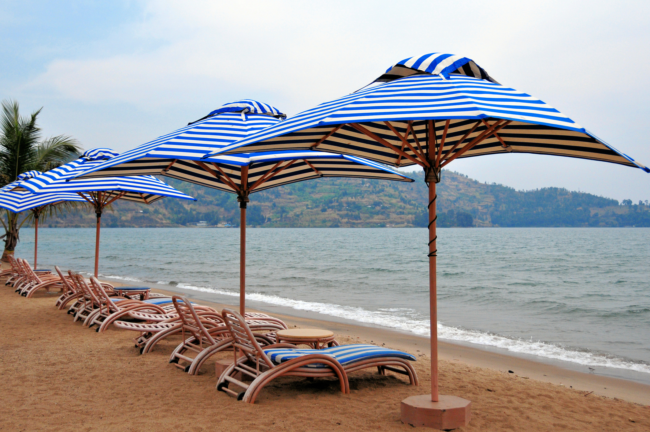 Row of blue and white striped beach umbrellas on sandy shore, with lounge chairs and calm sea in the background.