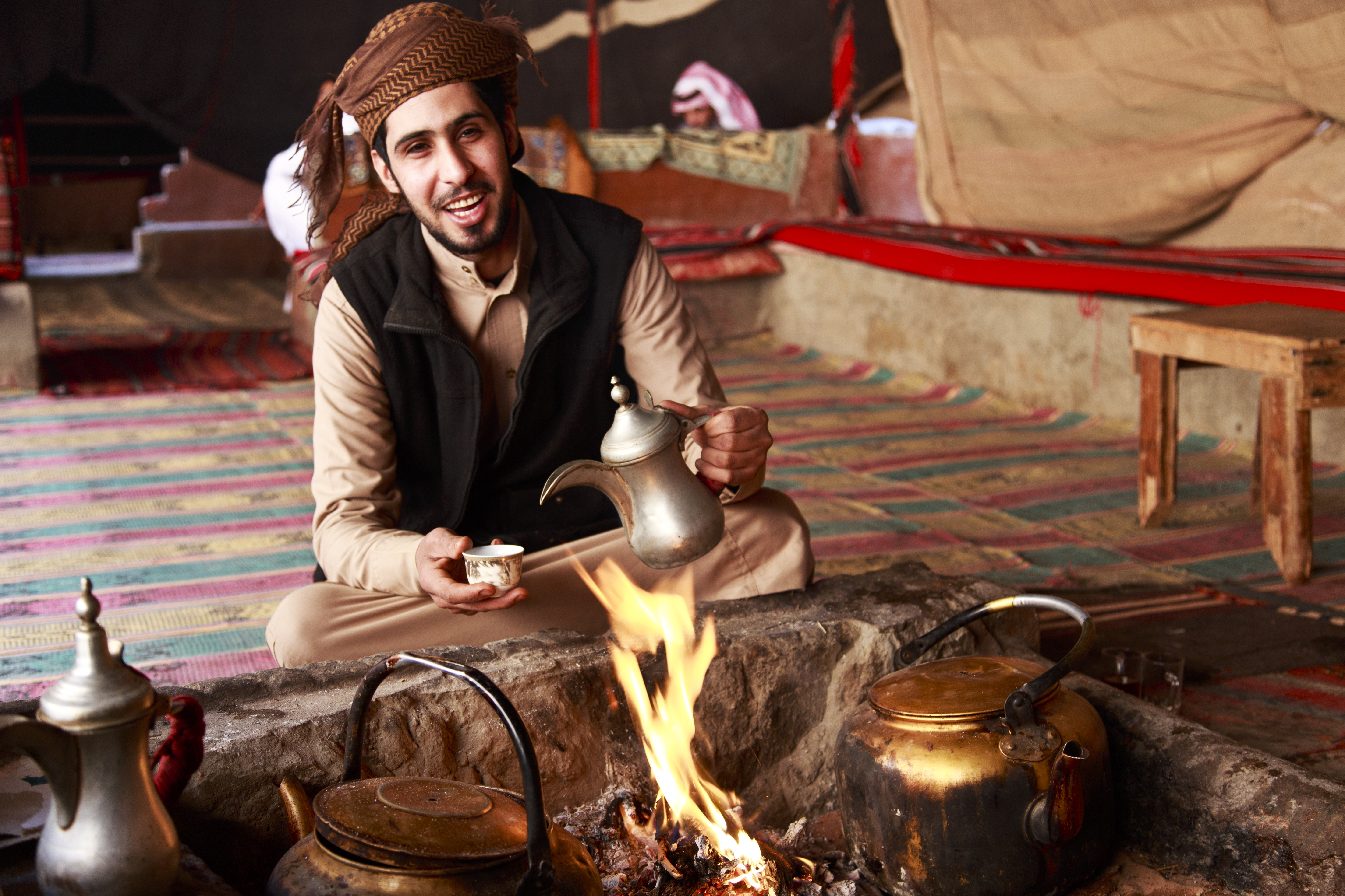A man in traditional attire sits by a fire, pouring tea from a kettle in a cozy, tent-like setting.