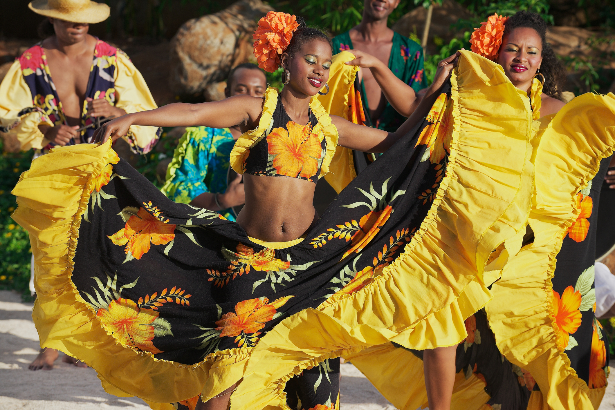 Two women in vibrant yellow and black dresses dance joyfully, surrounded by musicians dressed in colorful attire.