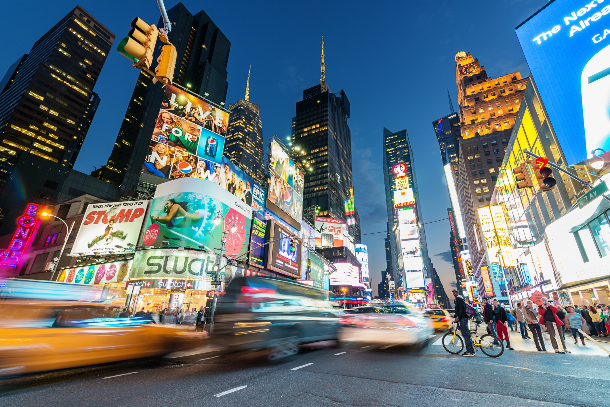 Brightly lit Times Square at dusk, with bustling traffic, pedestrians, and iconic billboards.