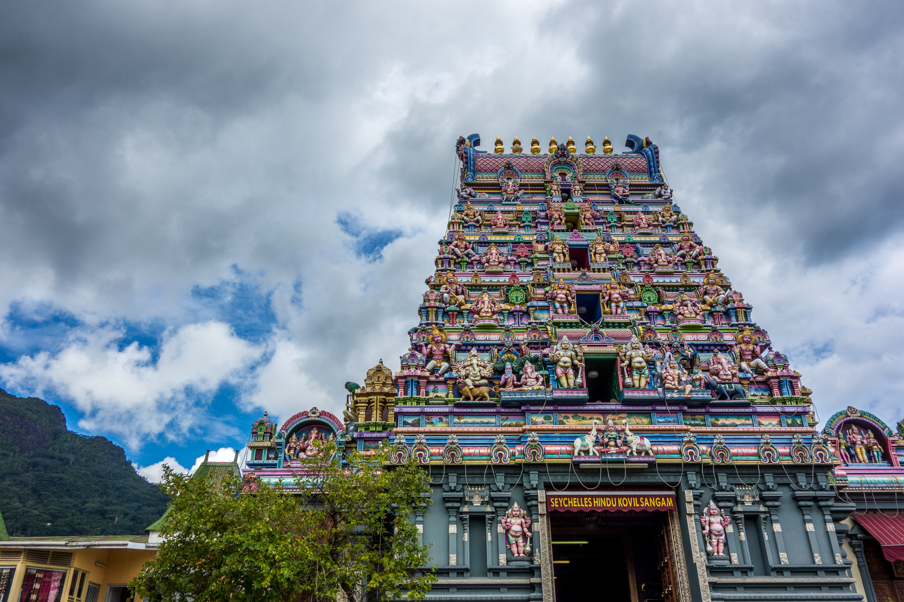 Colorful temple tower with intricate sculptures, set against a cloudy sky and lush greenery below.