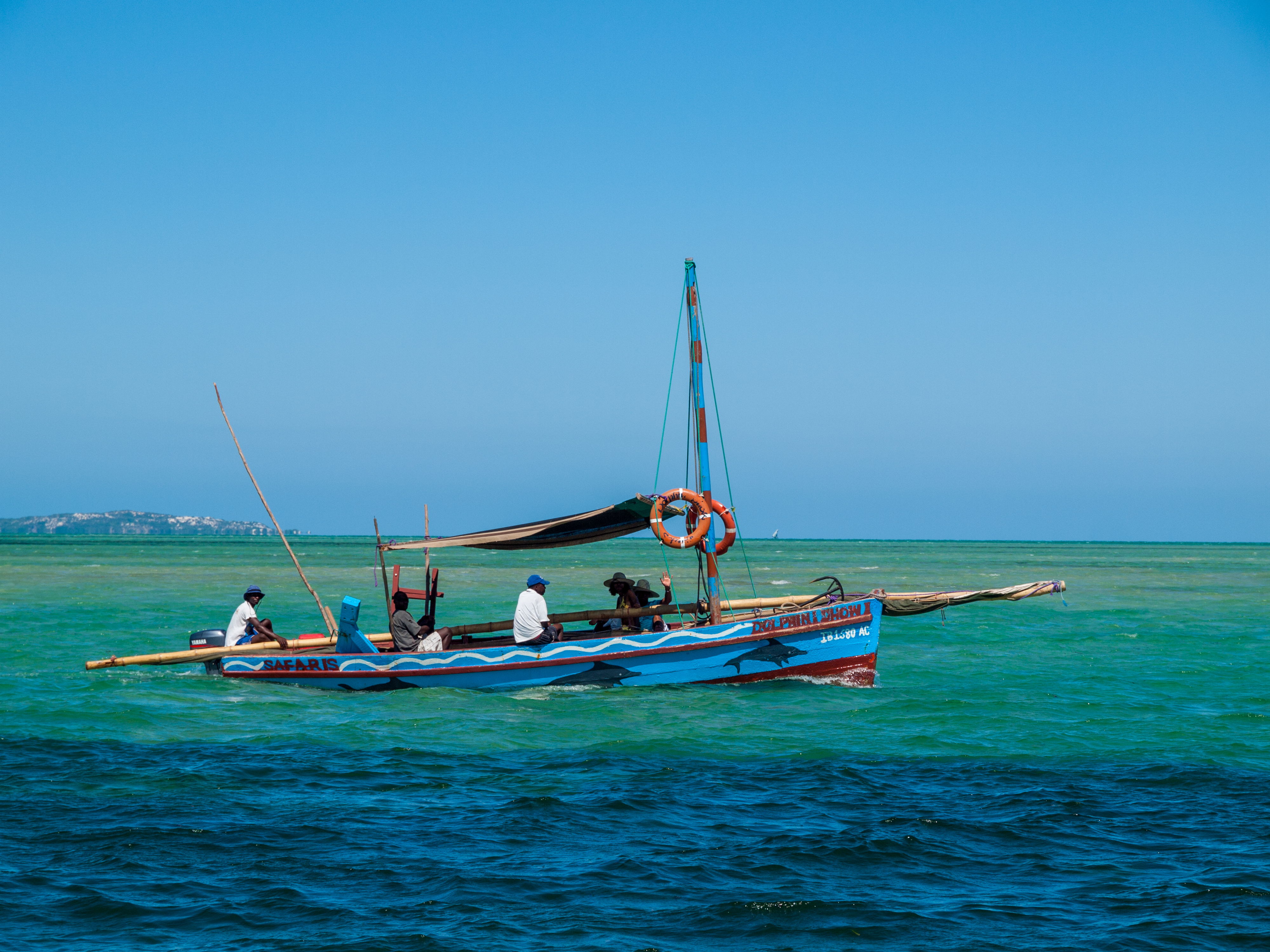 A colorful fishing boat with several people on the water, surrounded by a bright blue sky and clear green sea.