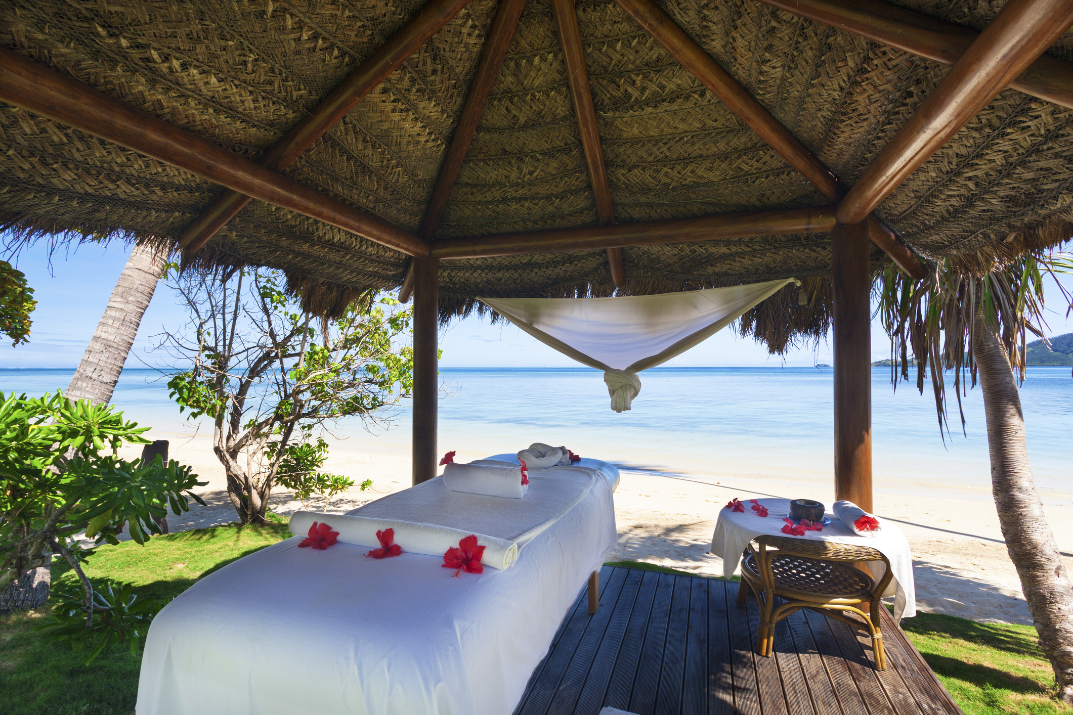 Spa treatment area with massage tables under a thatched roof, overlooking a serene beach and clear blue water.