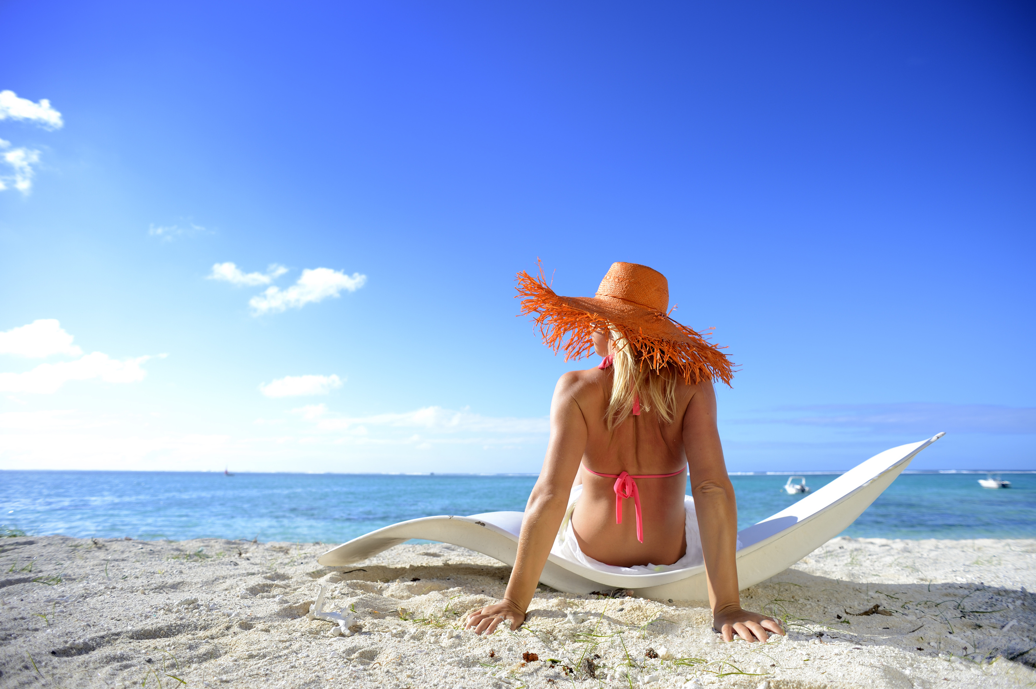 A person sits on the beach, wearing a large straw hat and a pink bikini, facing the ocean under a clear blue sky.
