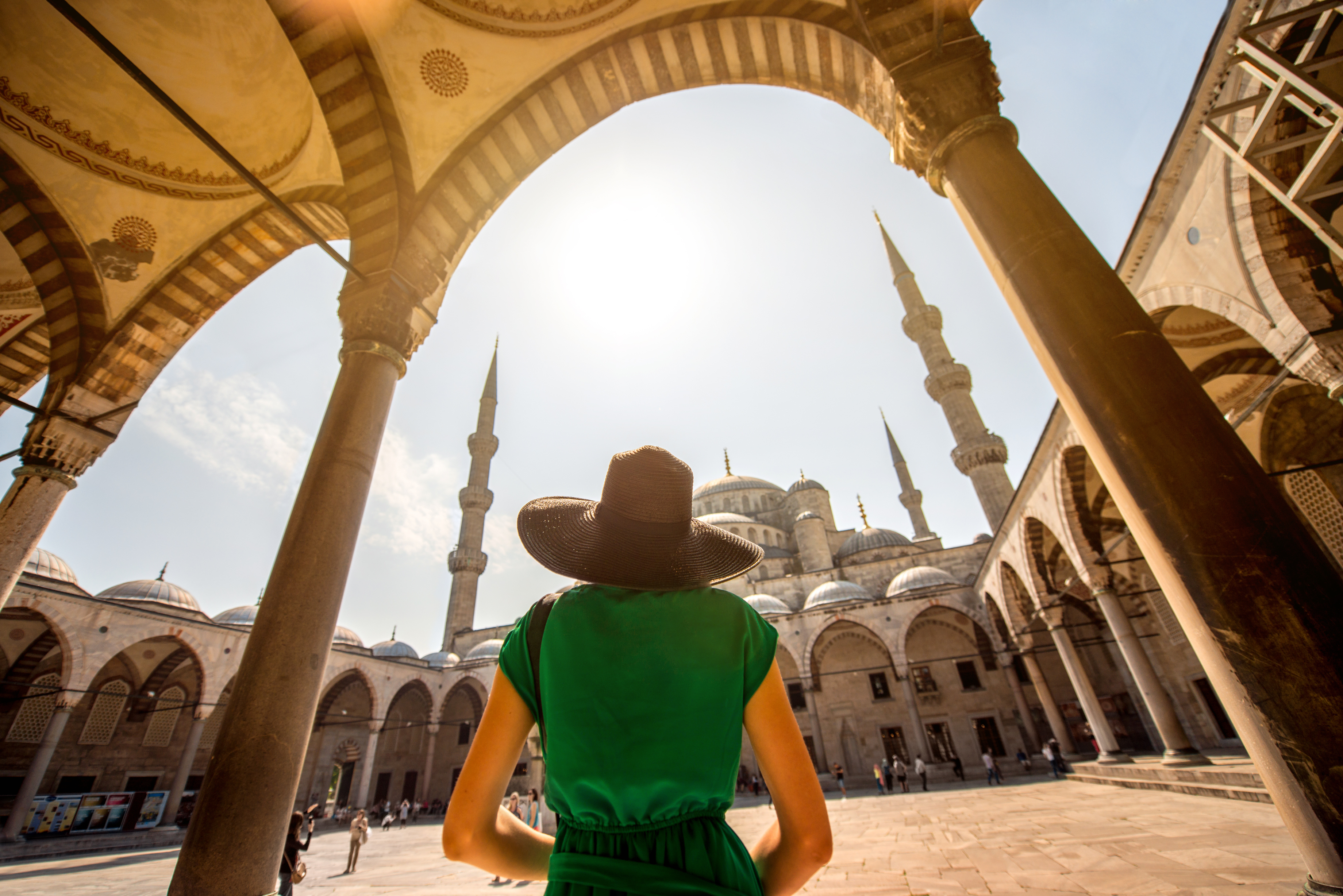 Person in a green dress and wide-brimmed hat stands under arches, overlooking a mosque with tall minarets and a bright sky.