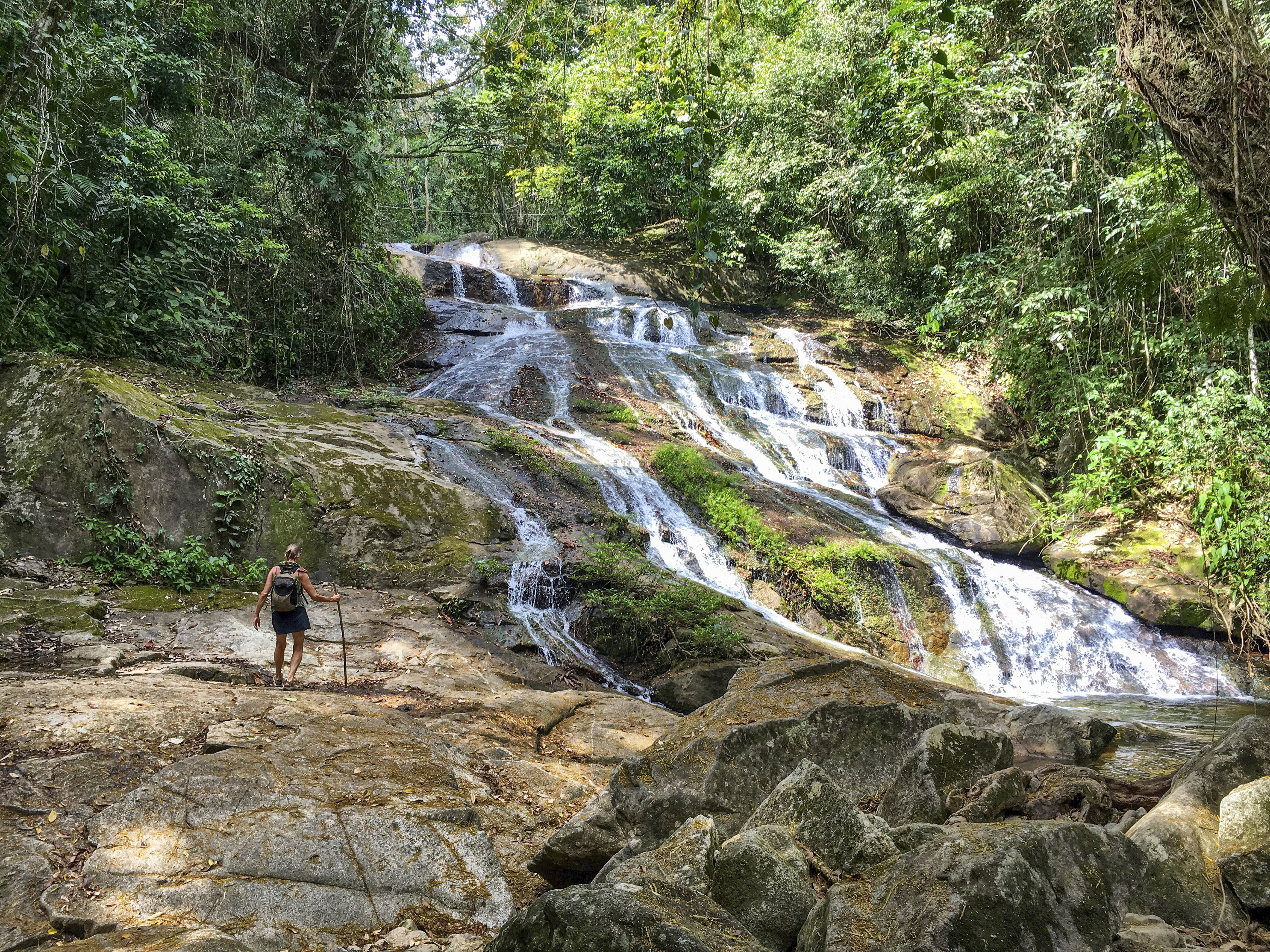 A hiker stands on rocky terrain near a cascading waterfall surrounded by lush greenery.