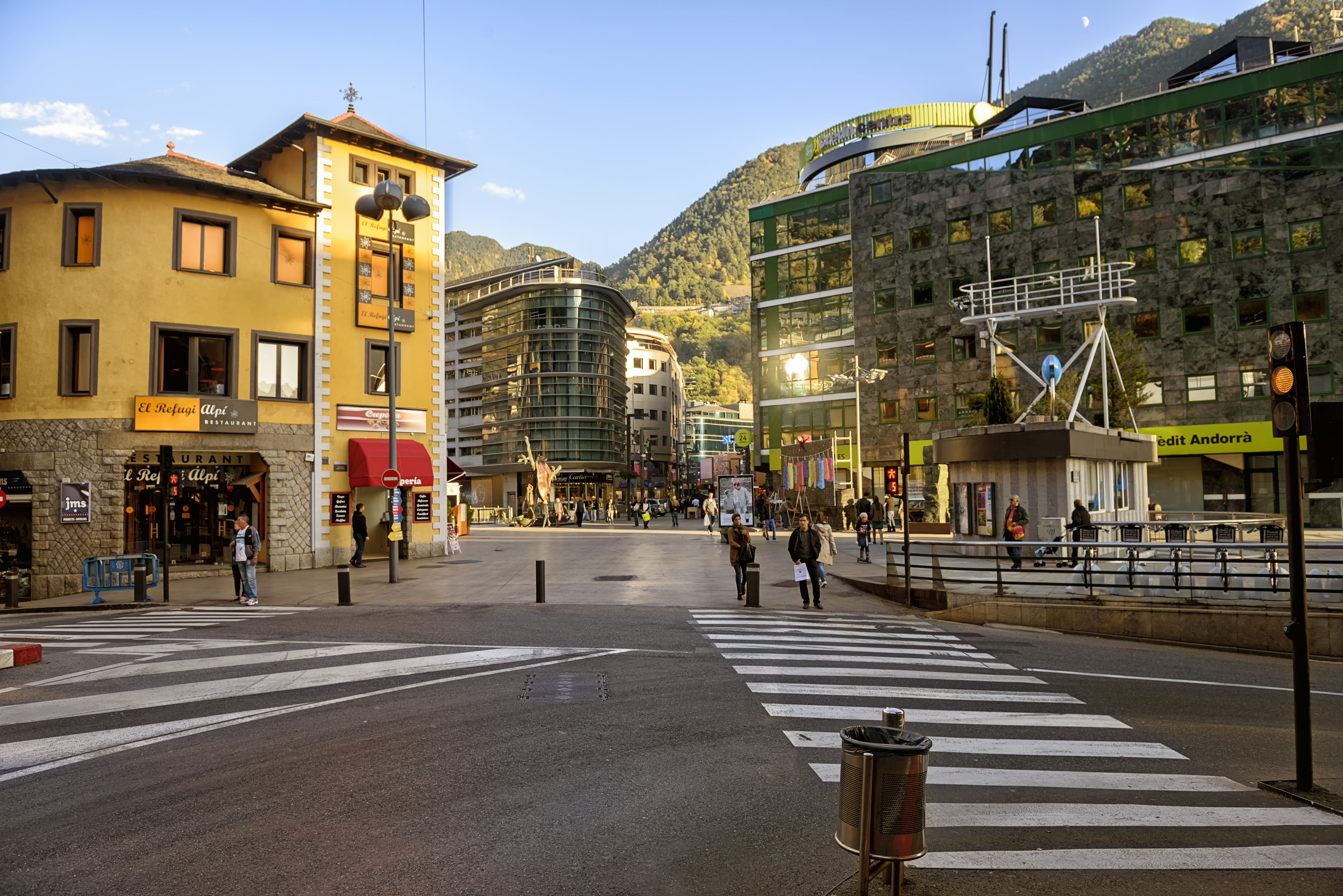 A city square with a mix of modern and traditional buildings, pedestrians crossing a striped crosswalk under a clear sky.
