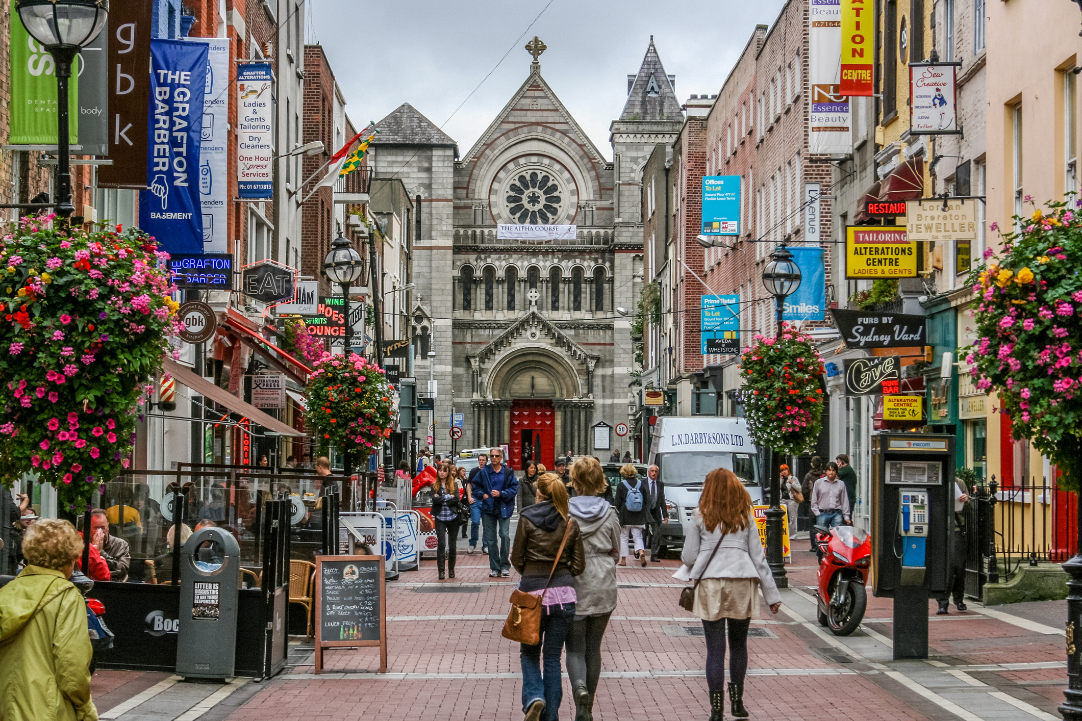 Bustling city street lined with shops and colorful flowers, leading to a historic church in the background.