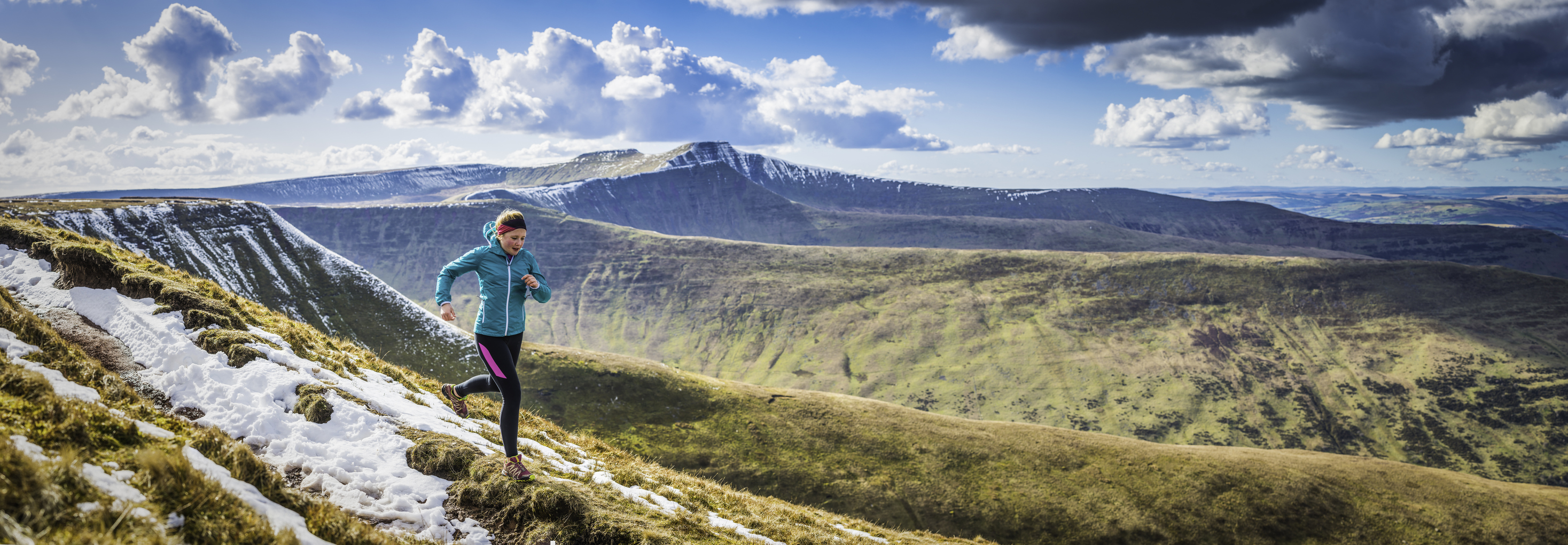 A person runs on a snowy mountain trail with scenic hills and clouds in the background.
