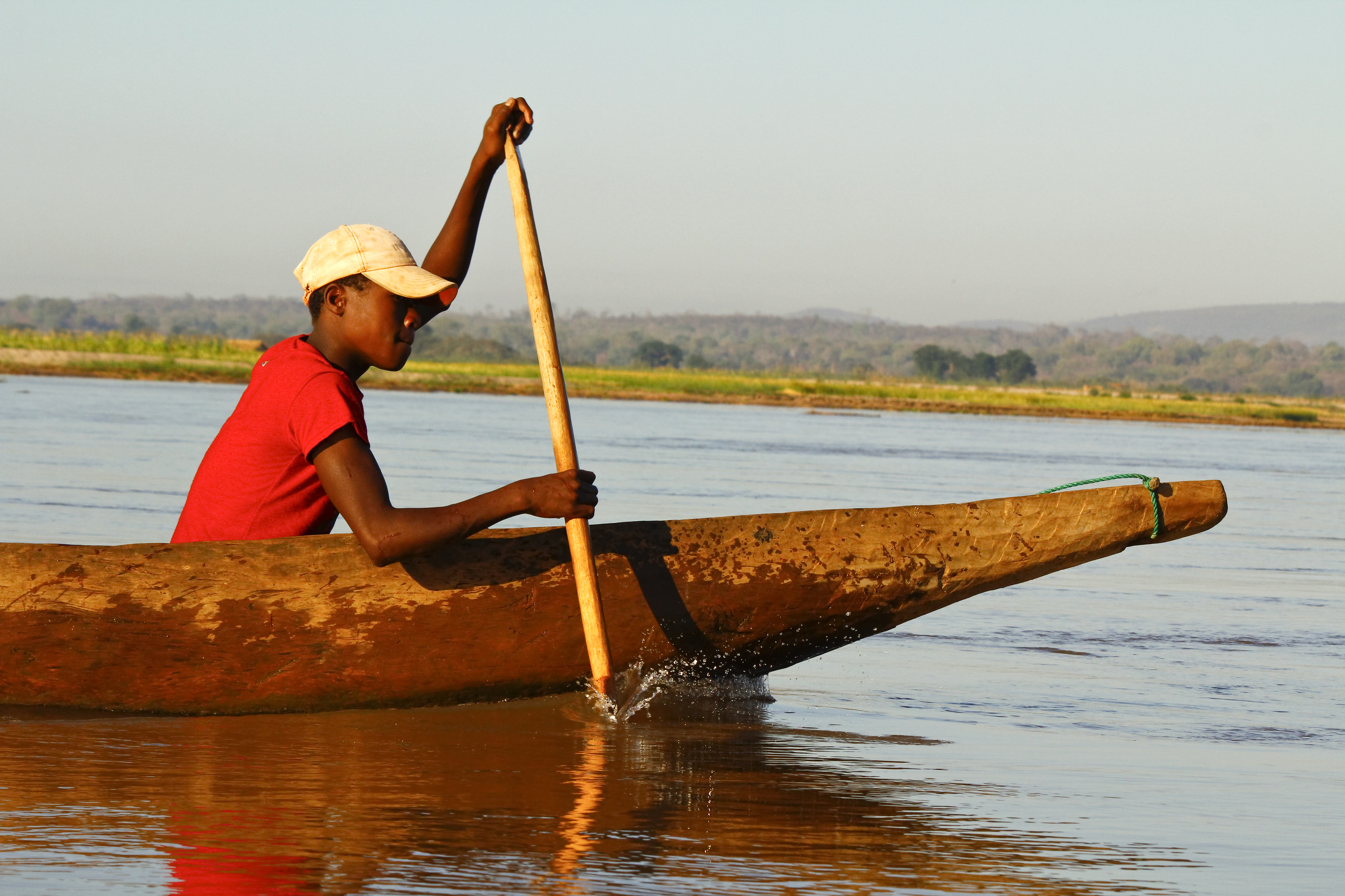 A person in a red shirt paddles a wooden canoe on a calm river during daylight.