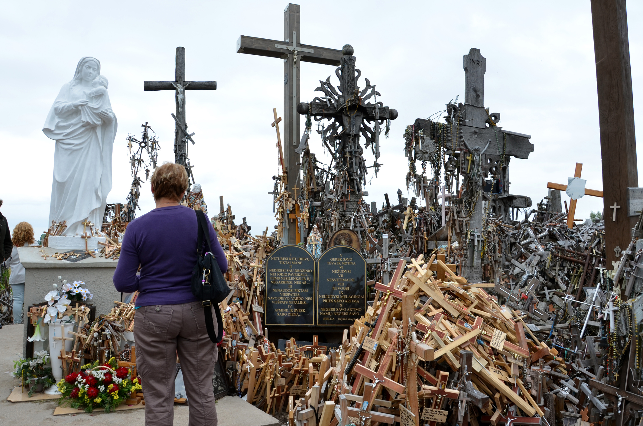 A woman stands before a shrine filled with wooden crosses and a statue, reflecting a memorial site.