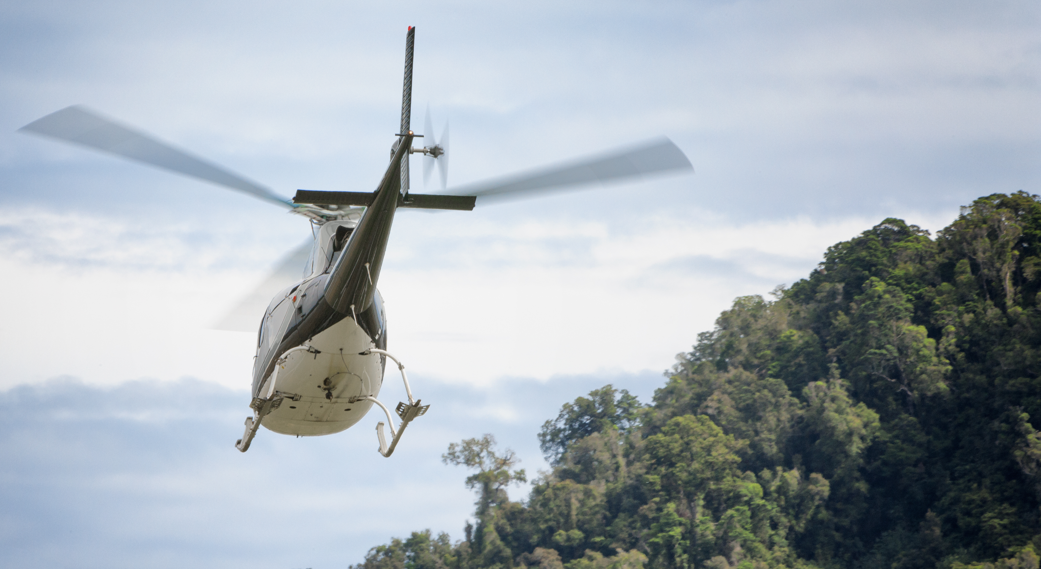 A helicopter flies above a lush green hillside under a cloudy sky.
