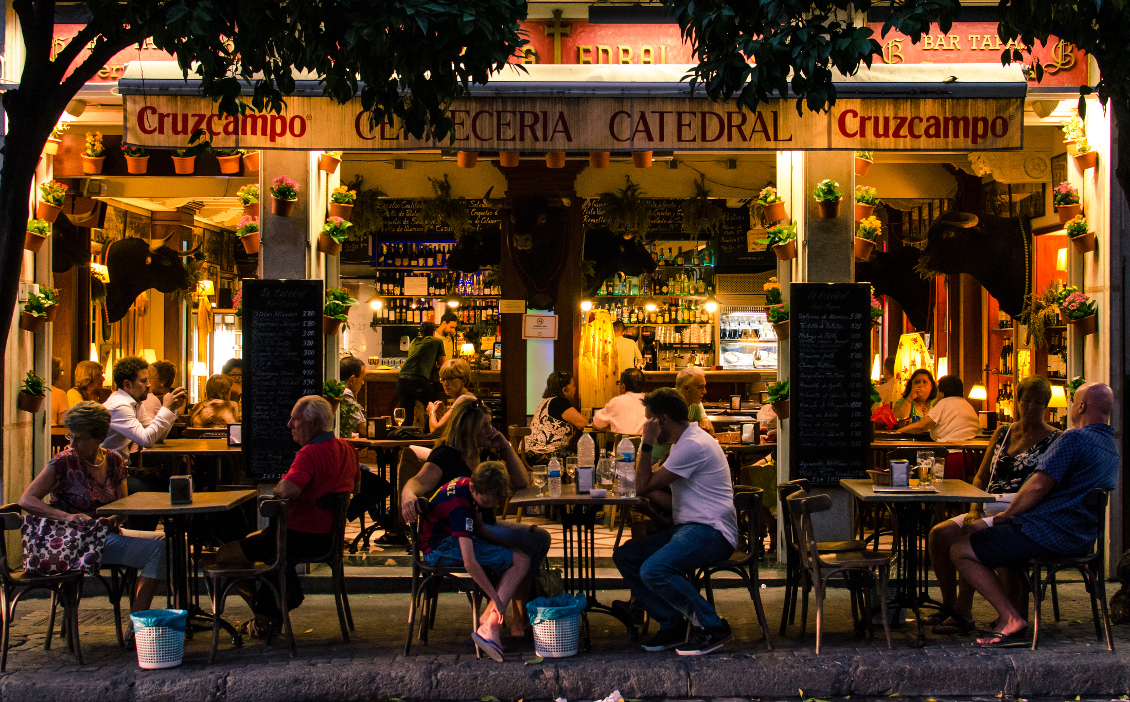A lively outdoor café with people dining at tables, illuminated by warm lights, surrounded by greenery and a welcoming atmosphere.