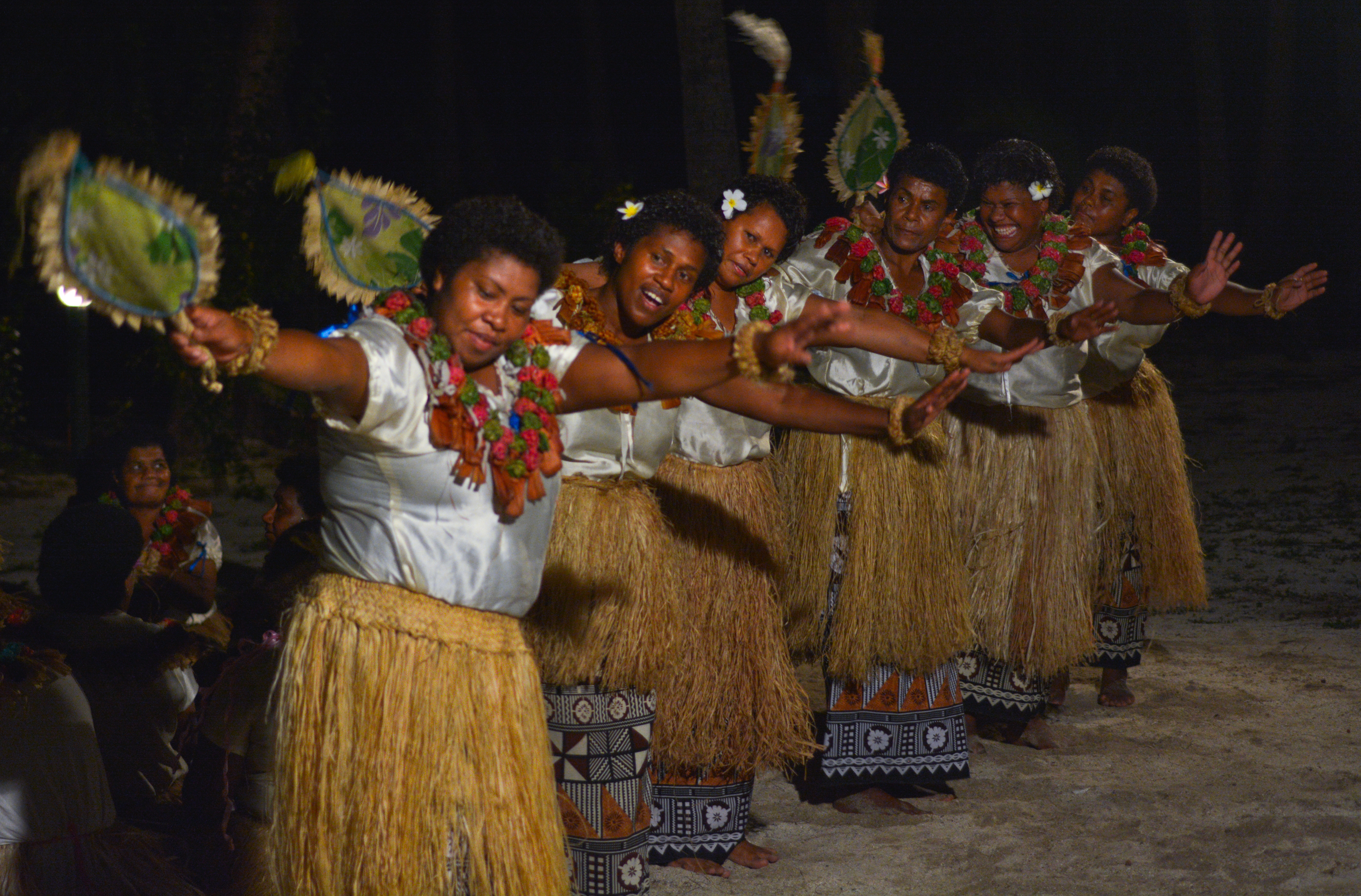 A group of women in traditional attire perform a dance with fans, showcasing vibrant cultural expression under a night sky.