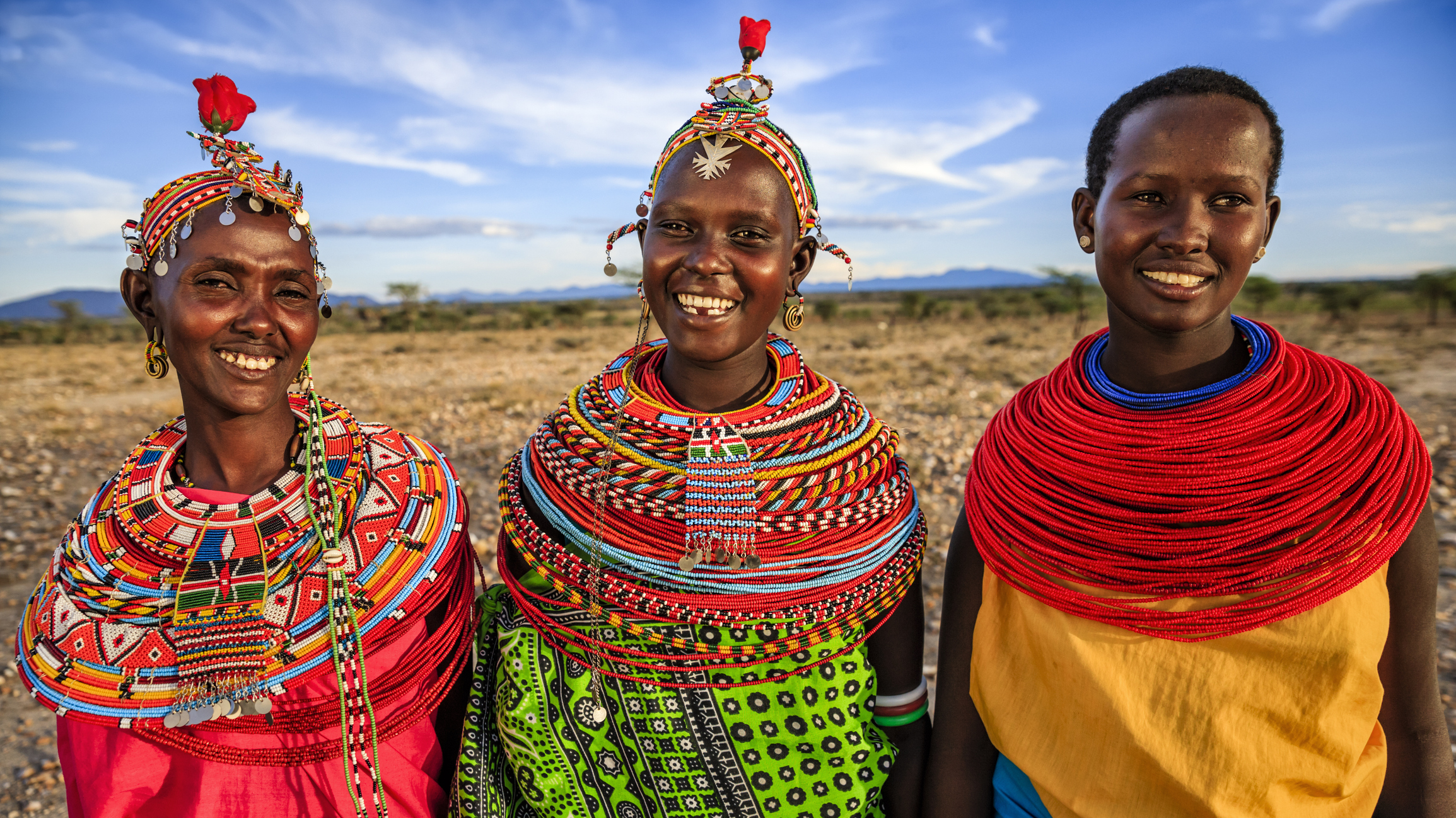Three smiling women in colorful traditional attire stand together against a scenic landscape.
