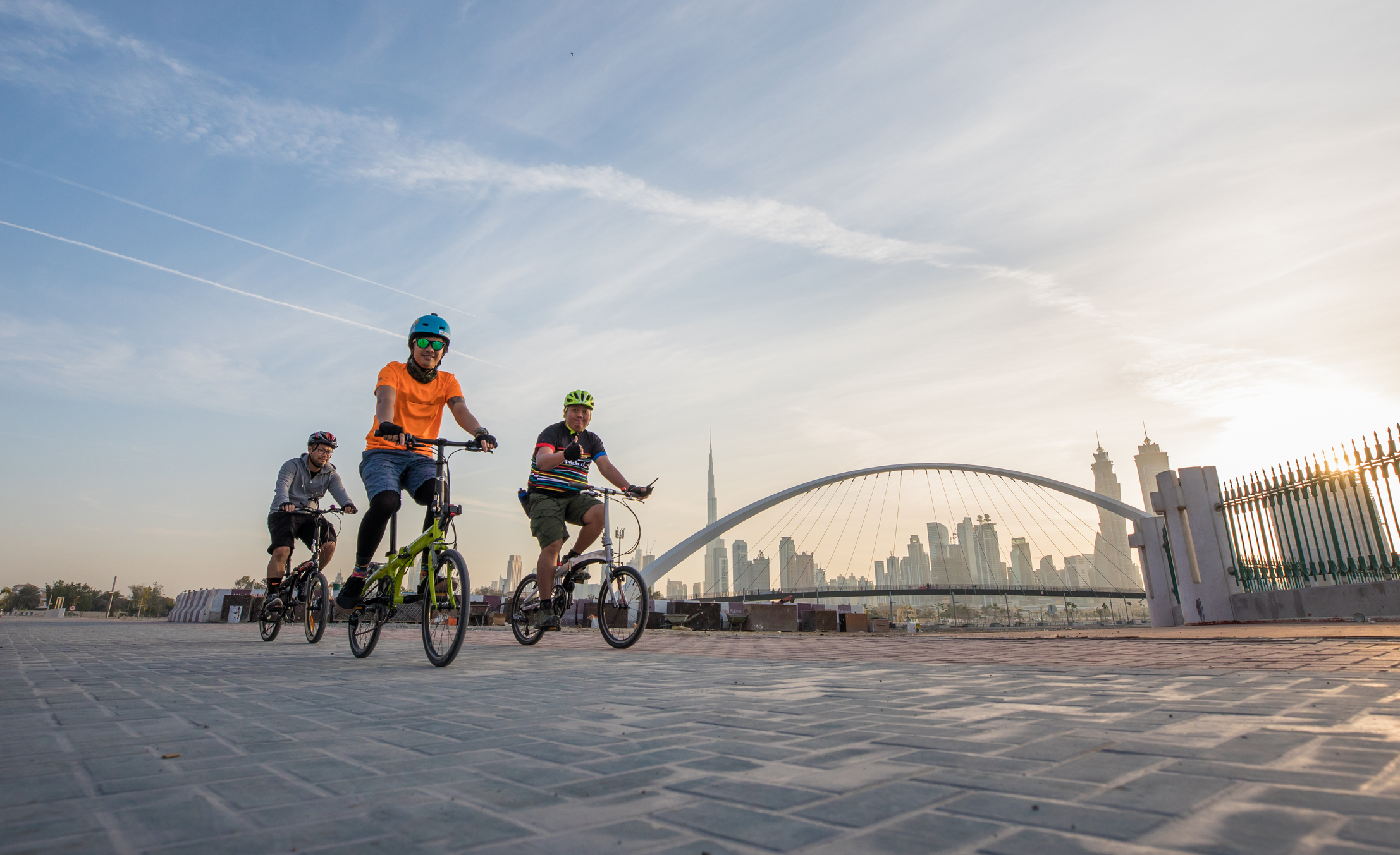 Three children ride bicycles along a path with a city skyline and bridge in the background under a sunset sky.