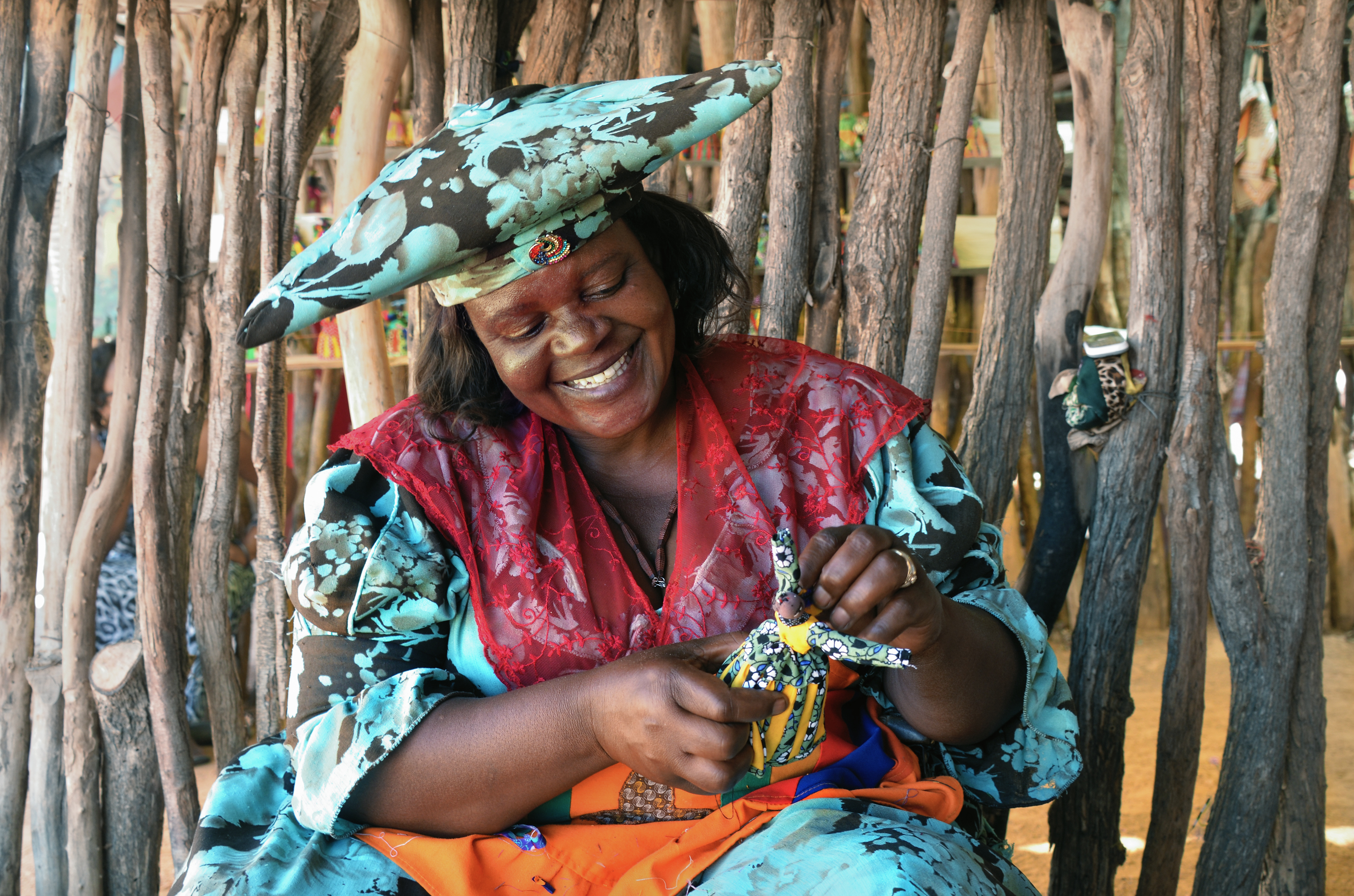 Smiling woman in colorful attire crafts a decorative doll, surrounded by natural wooden elements in a vibrant setting.