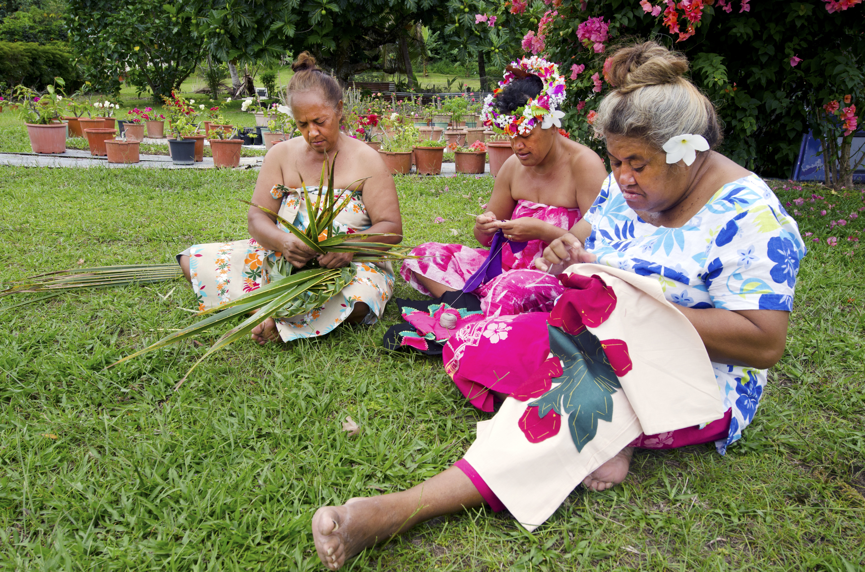 Three women sitting on grass, weaving with leaves and fabric, surrounded by colorful flowers and plants in pots.