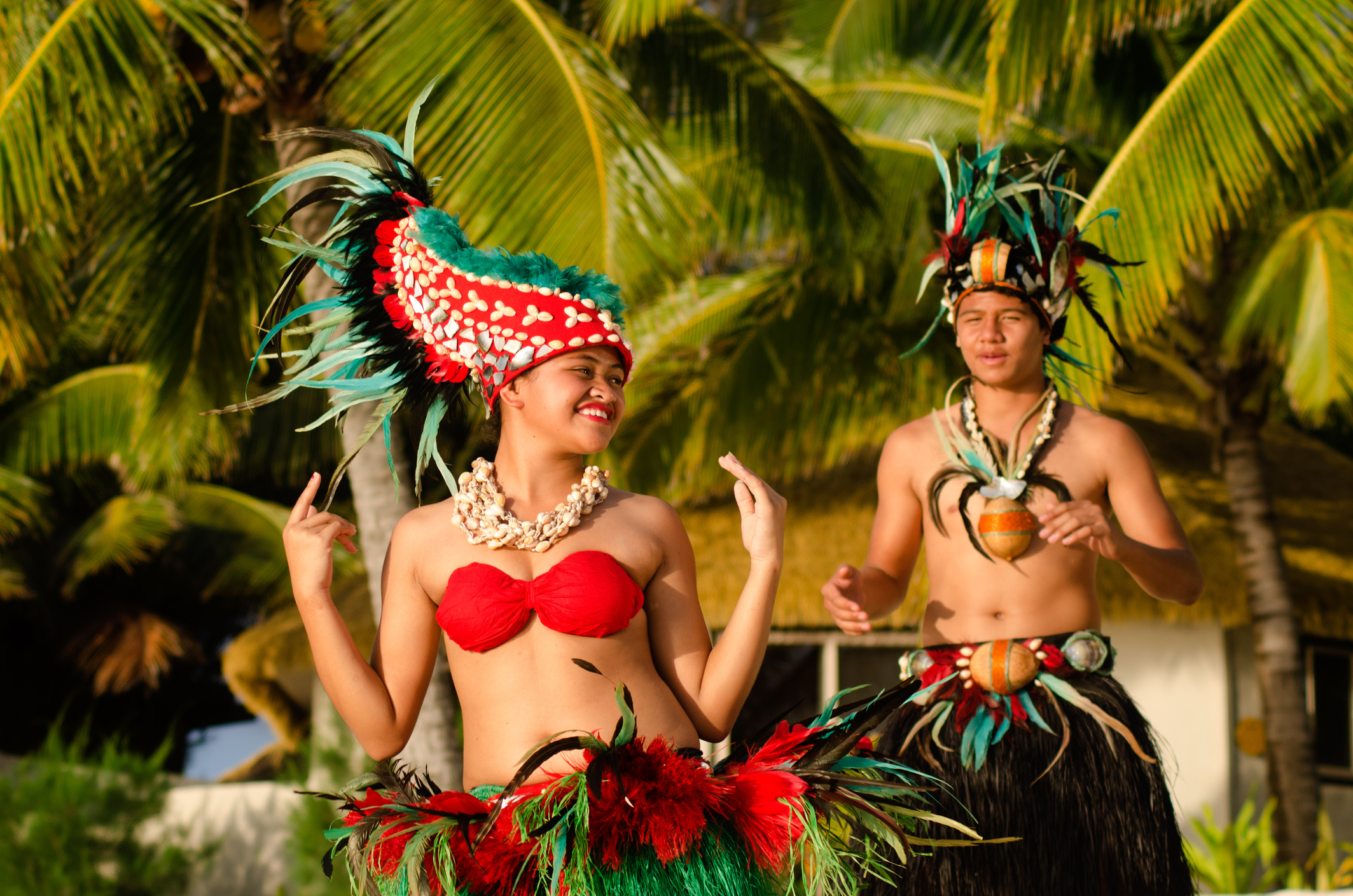Two dancers in traditional attire perform in a tropical setting, surrounded by palm trees and vibrant greenery.