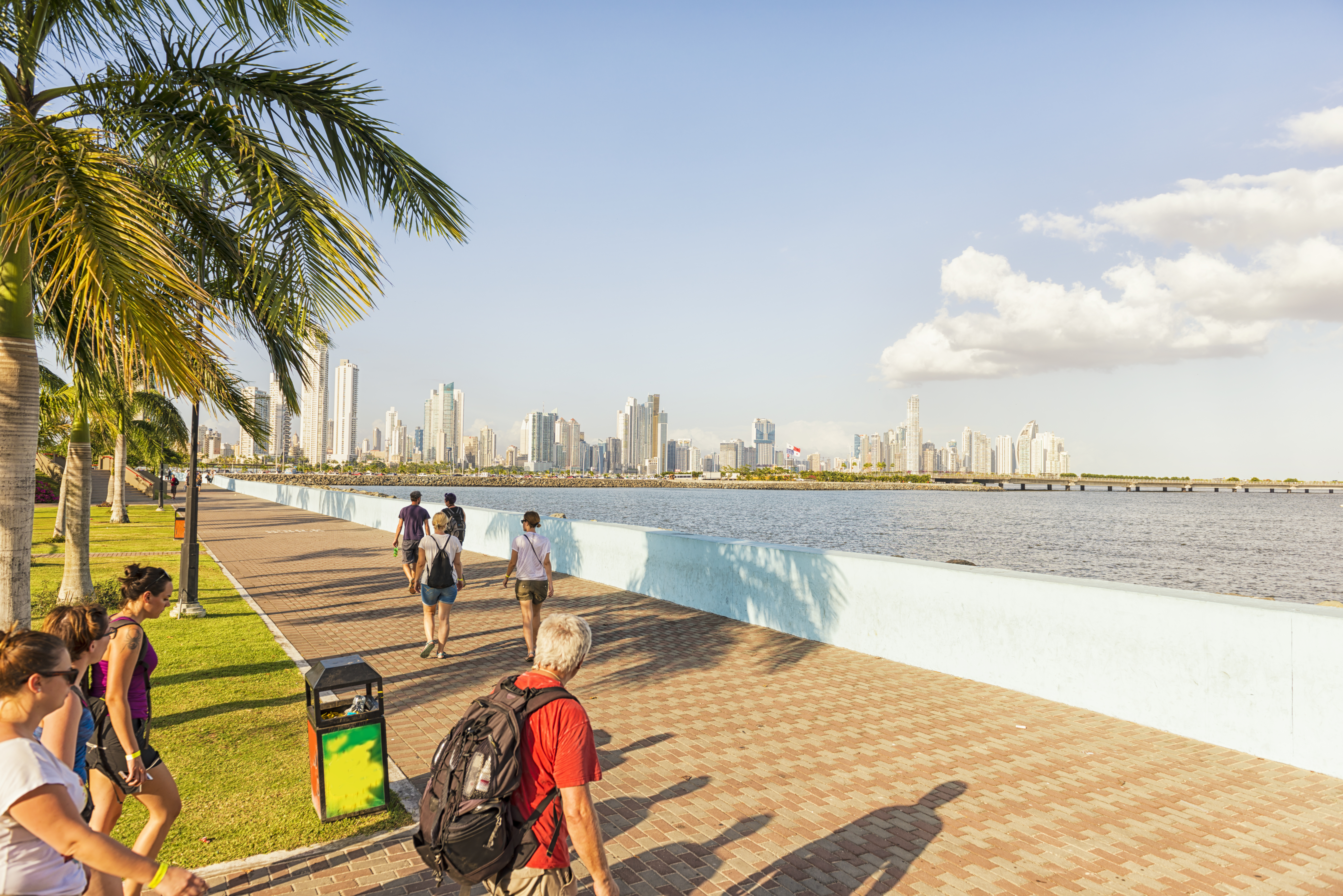 People walking along a waterfront promenade with a skyline and palm trees in the background under a clear sky.