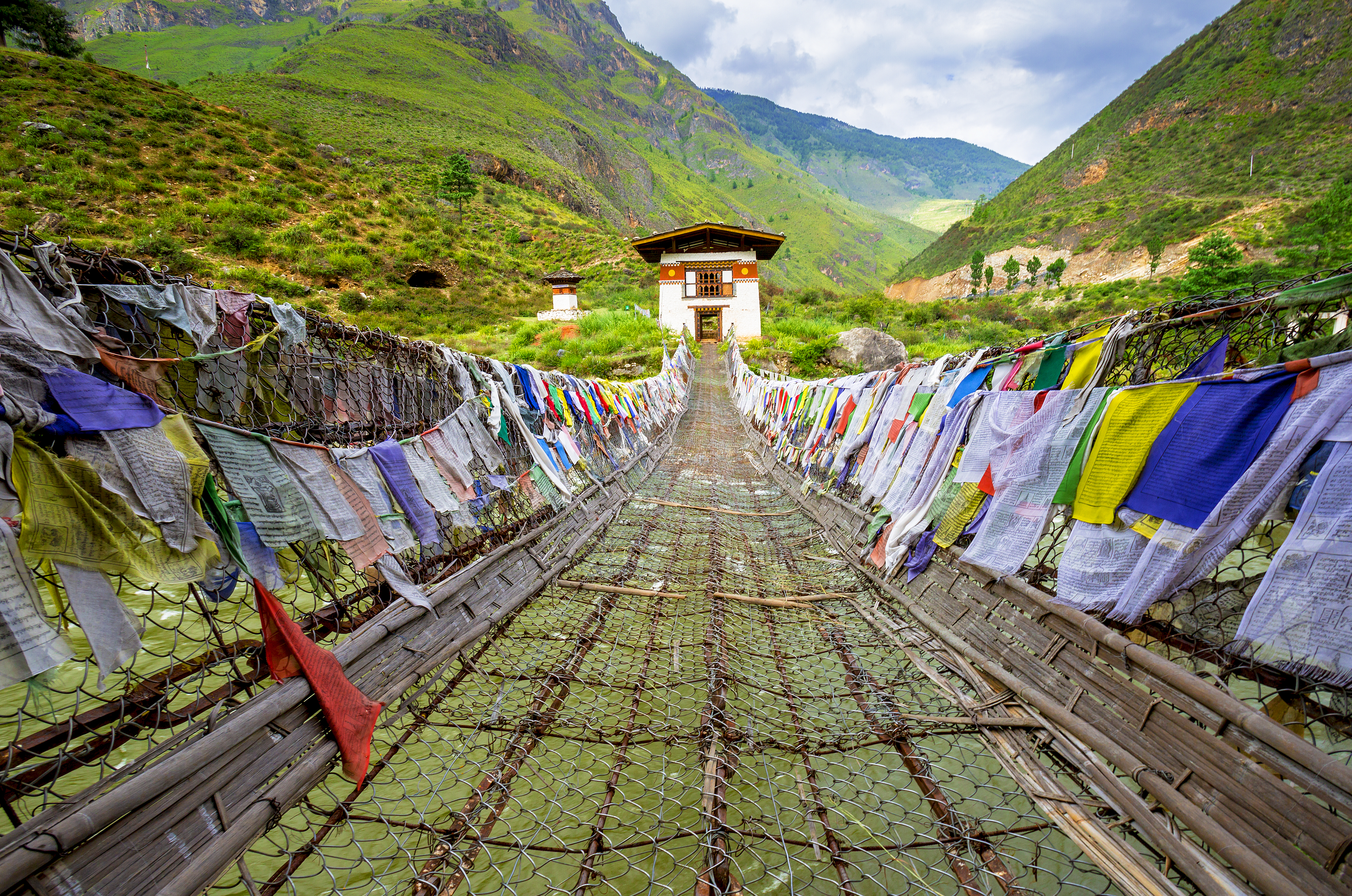 View straight on of Iron Chain Bridge of Tamchog Lhakhang Monastery with prayer flags lining the bridge, mountains and cloudy sky in the background