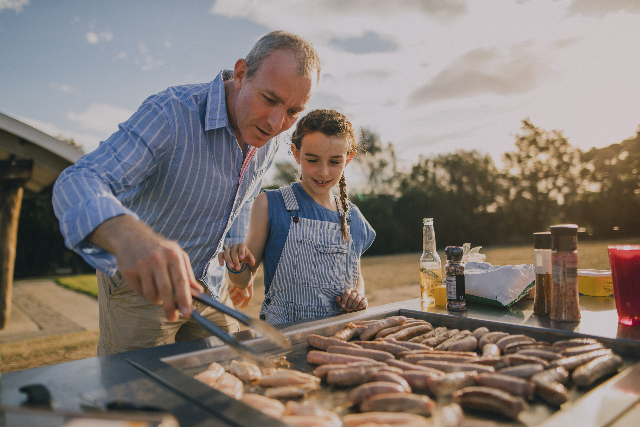 An adult man and a girl are grilling sausages together at a barbecue, enjoying the outdoor setting.