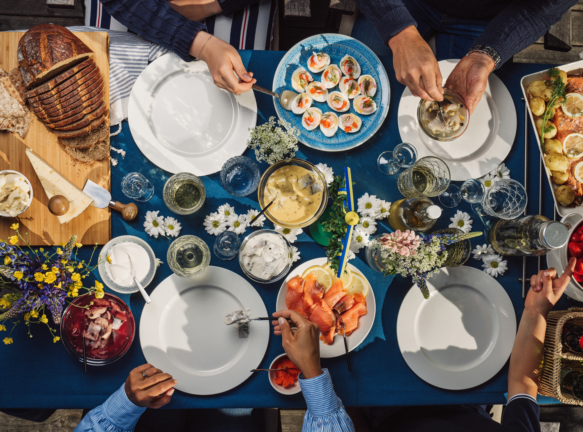 Aerial view of a vibrant outdoor table set with various dishes, drinks, and floral decorations, with hands reaching for food.