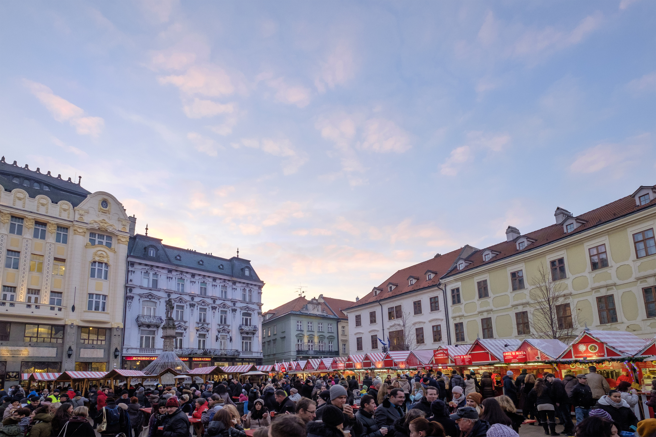 Crowded market scene with stalls under a colorful sky, surrounded by historic buildings.