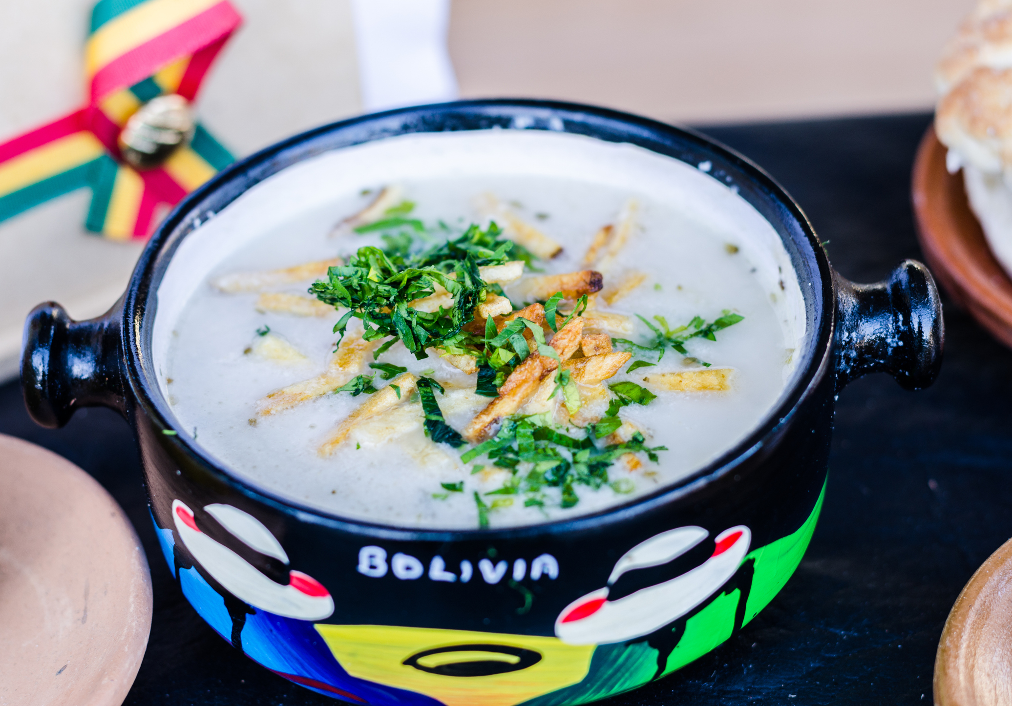 A colorful bowl of traditional Bolivian soup topped with herbs and crispy strips, alongside bread on a wooden surface.