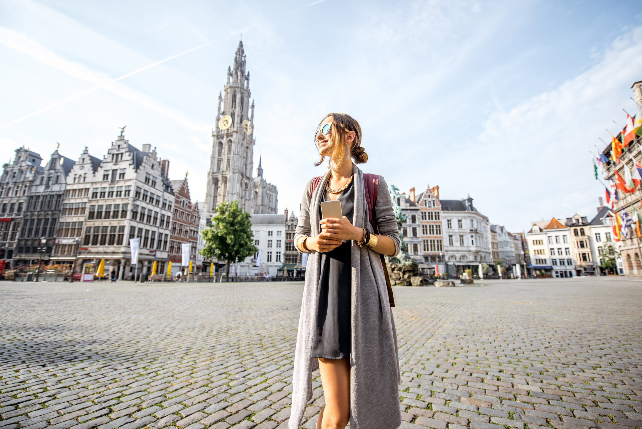 A woman stands in a plaza with historic buildings and a clock tower in the background, enjoying a sunny day.