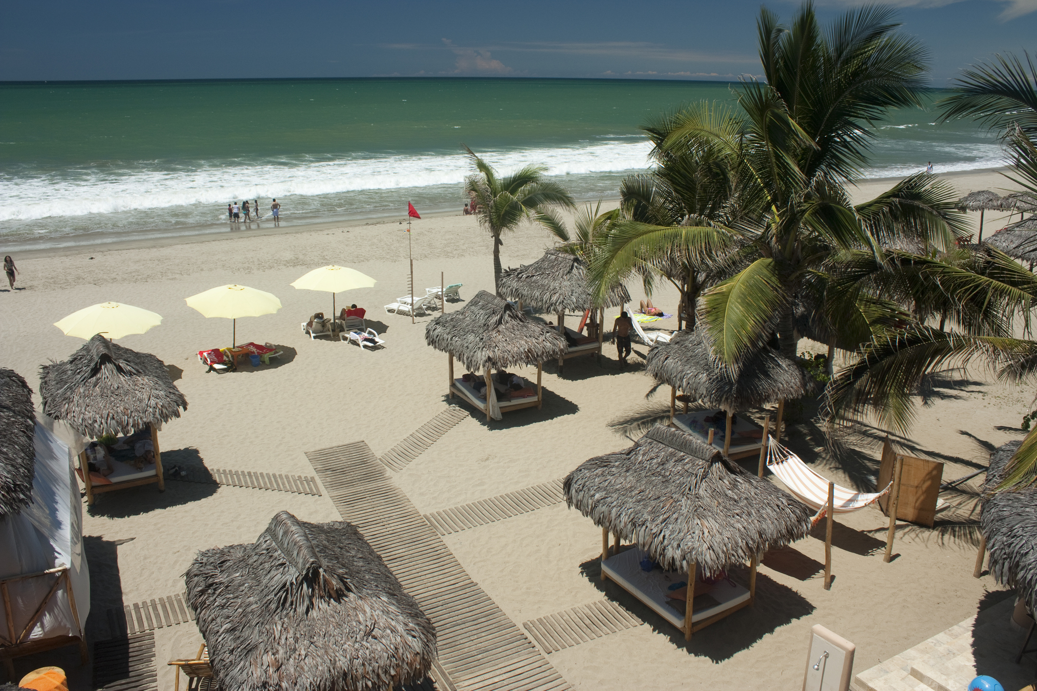 A beach scene with straw huts, palm trees, and umbrellas, featuring gentle waves and people in the background.