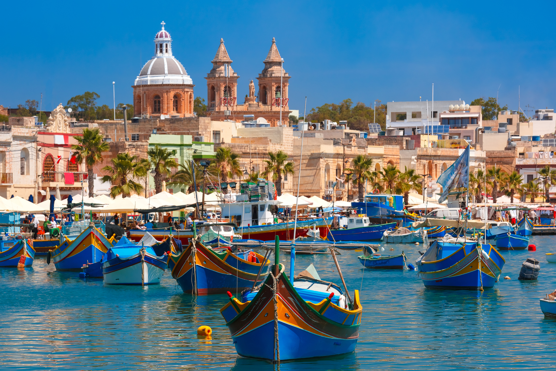 Colorful boats in a vibrant harbor, with historic buildings and palm trees under a bright blue sky.