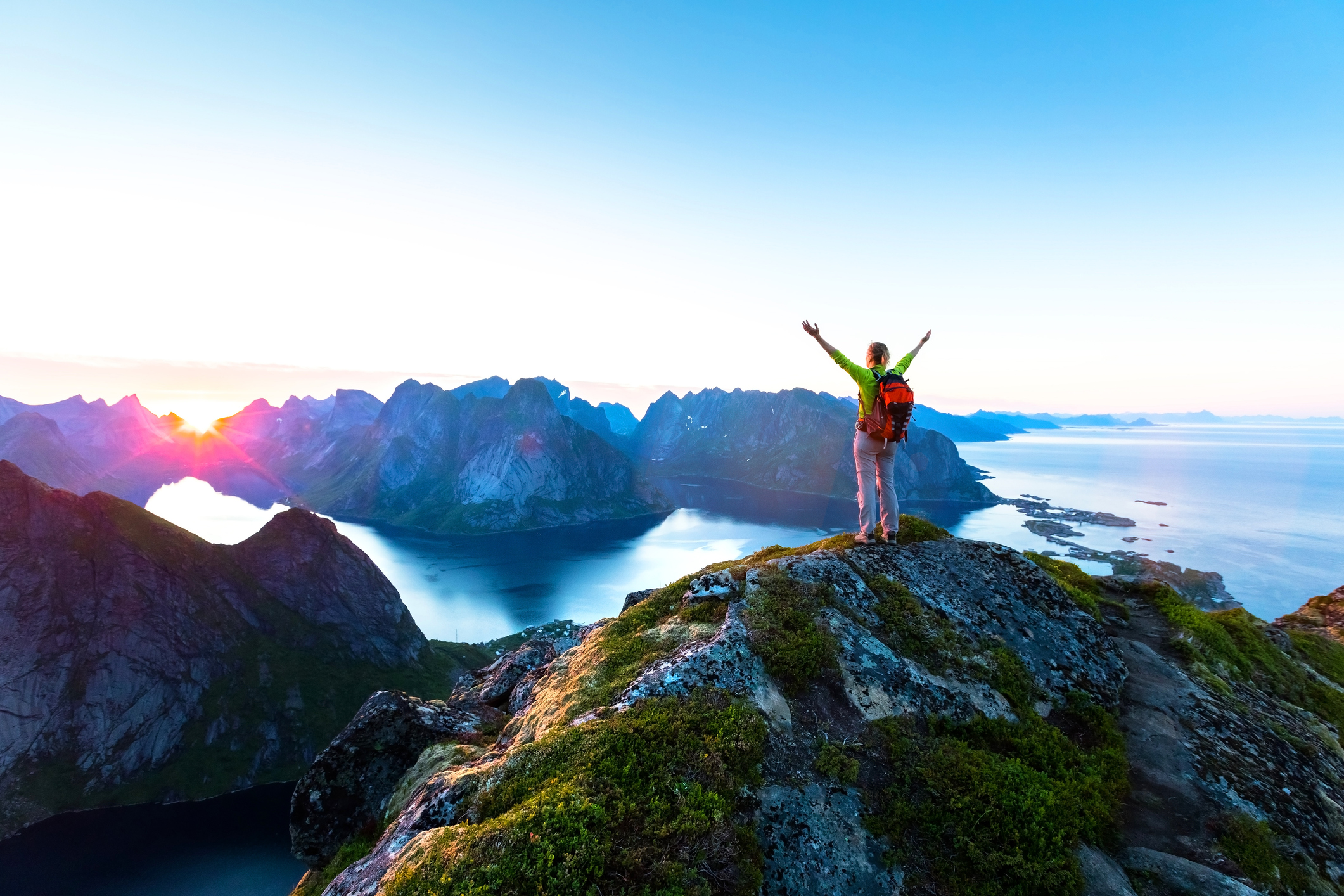 A person with arms raised stands on a rocky peak, overlooking a colorful landscape of mountains and water at sunrise.
