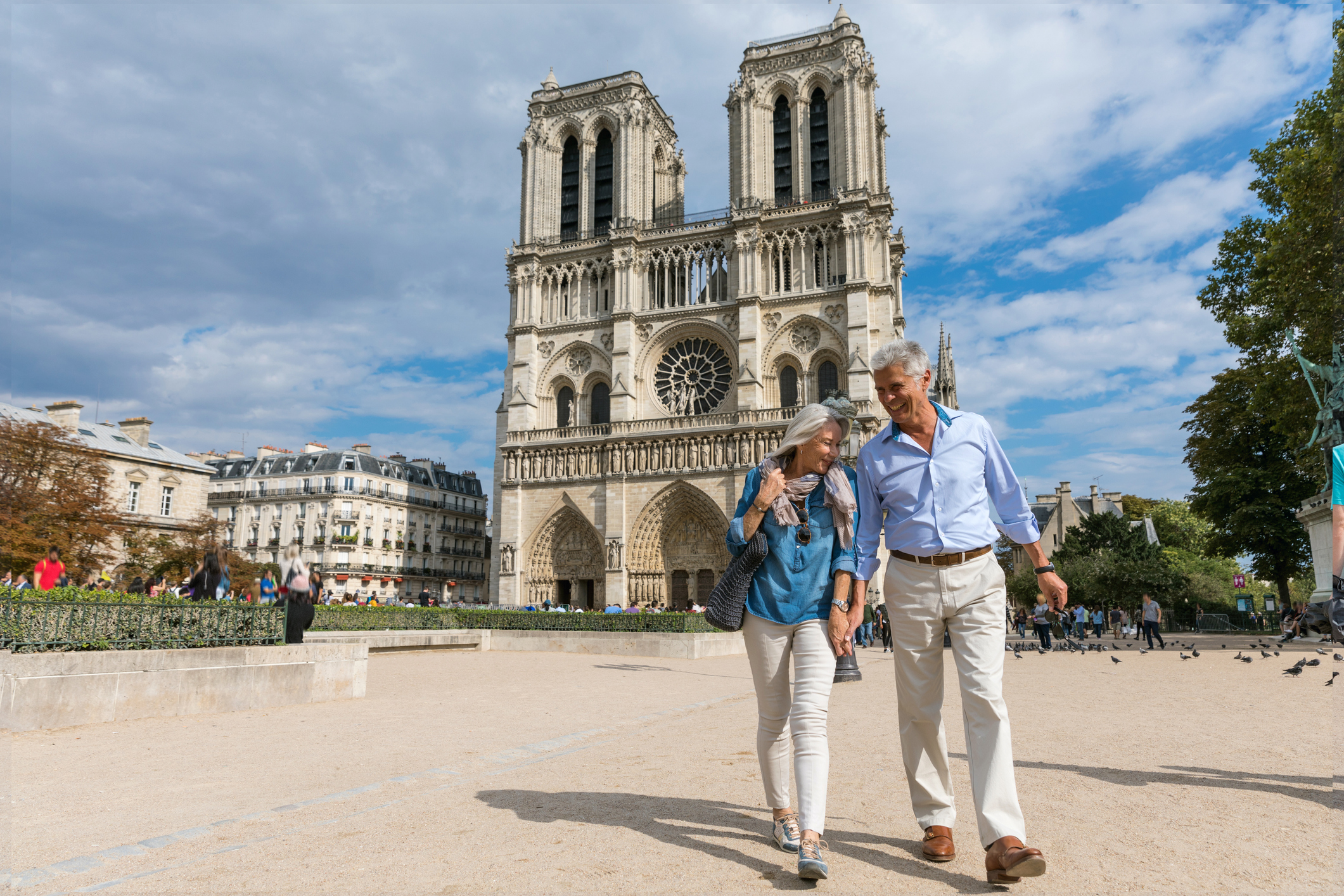 A couple walks hand-in-hand in front of Notre-Dame Cathedral on a sunny day, with people and trees in the background.