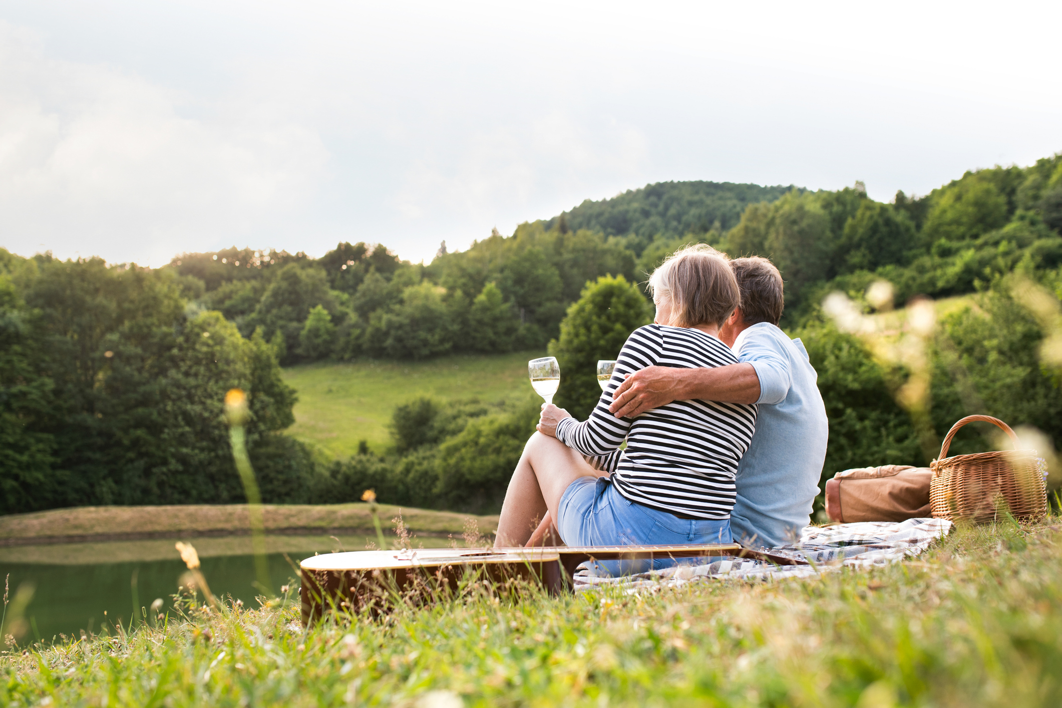A couple sits together by a serene lake, enjoying a drink and the view of green hills and trees in the background.