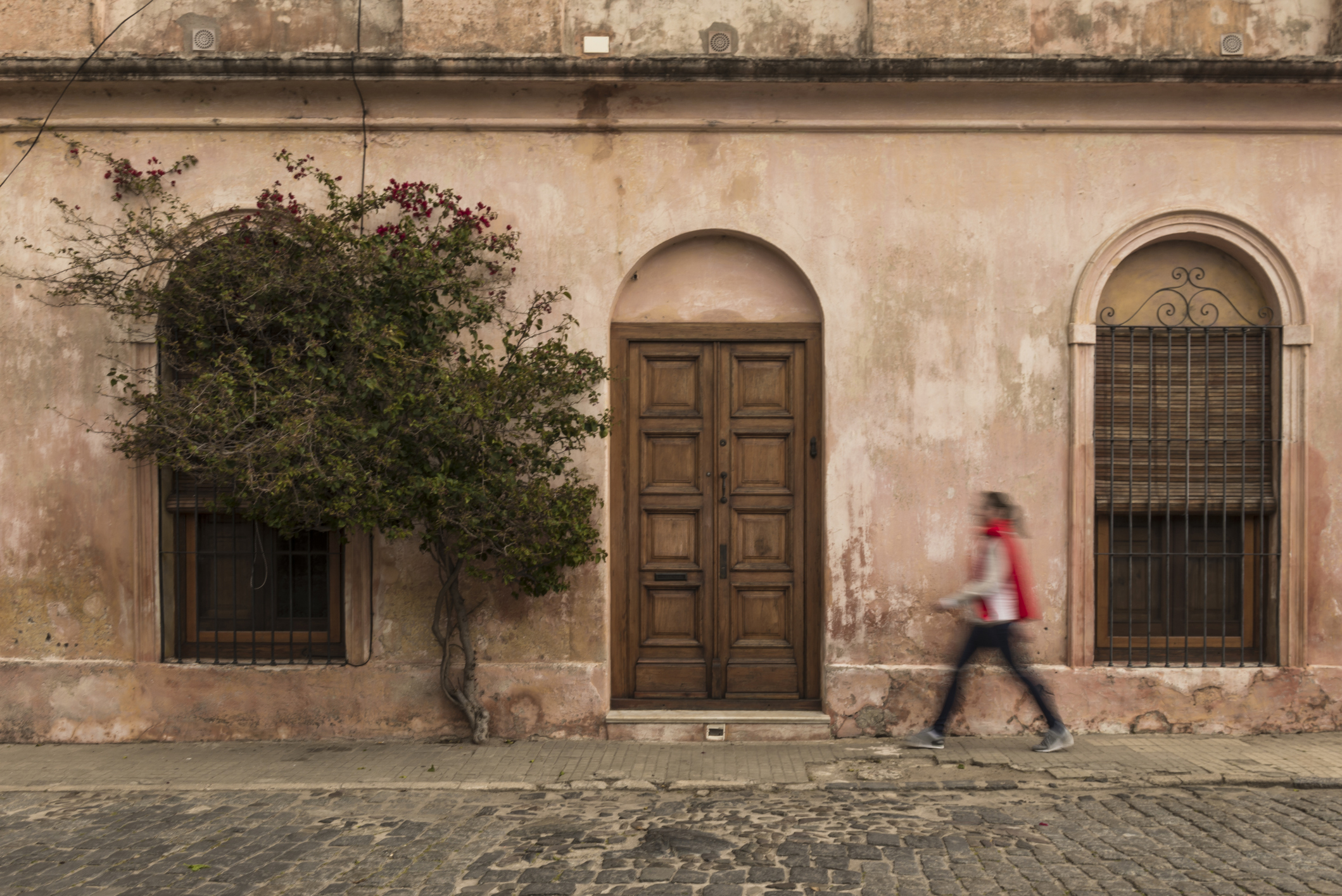 A person walks past a weathered pink building with an arched door and windows, accompanied by a small tree.