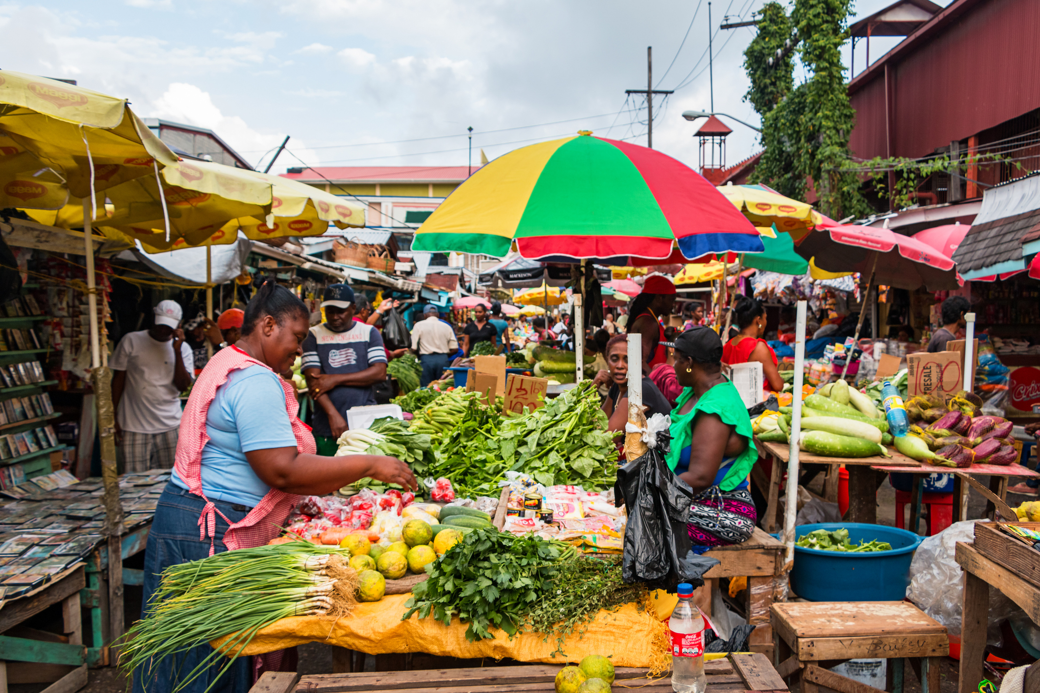 A vibrant market scene featuring vendors selling fresh produce under colorful umbrellas, with shoppers browsing the stalls.