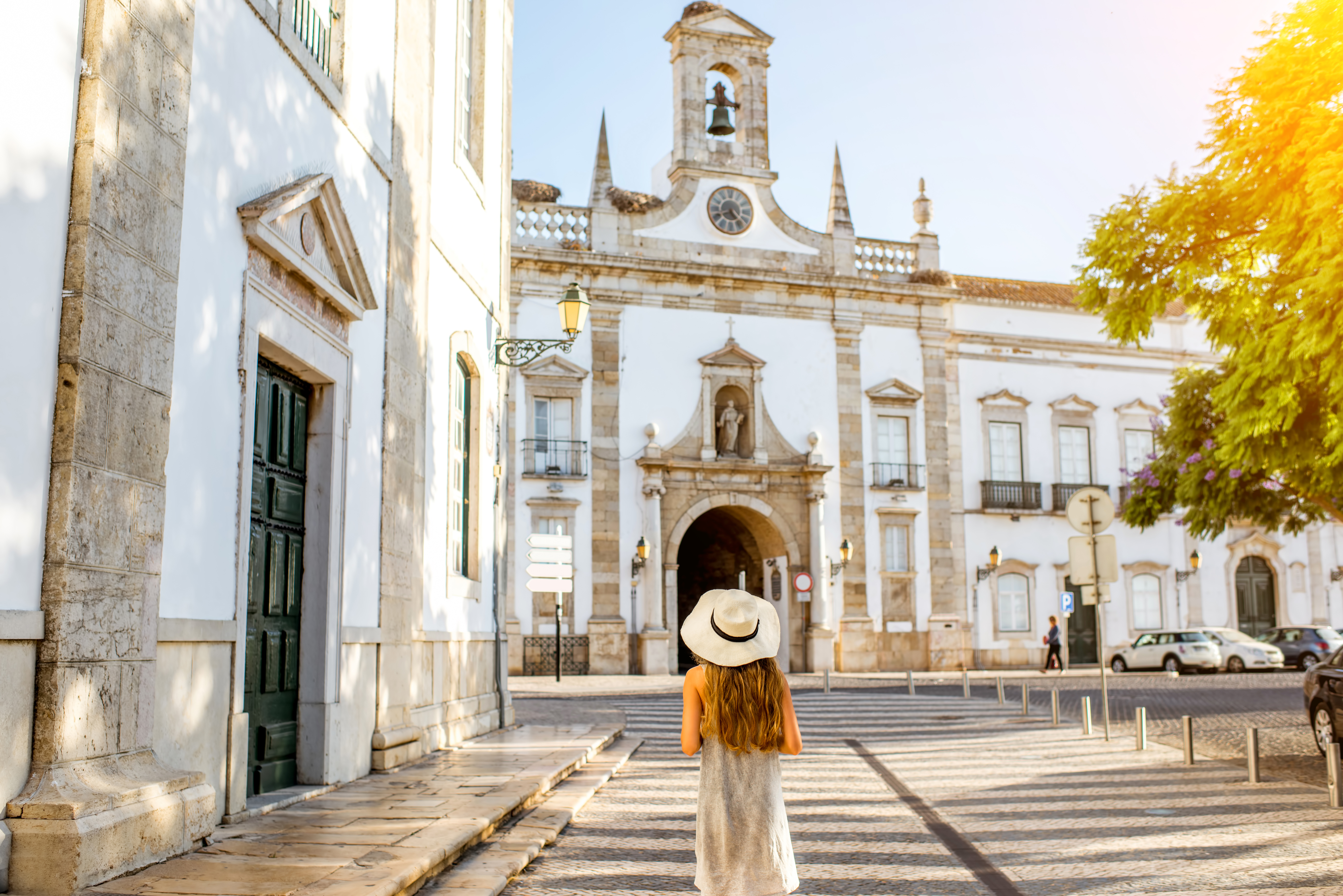 A woman in a hat walks towards a historic building, surrounded by trees and sunlight in a charming street setting.