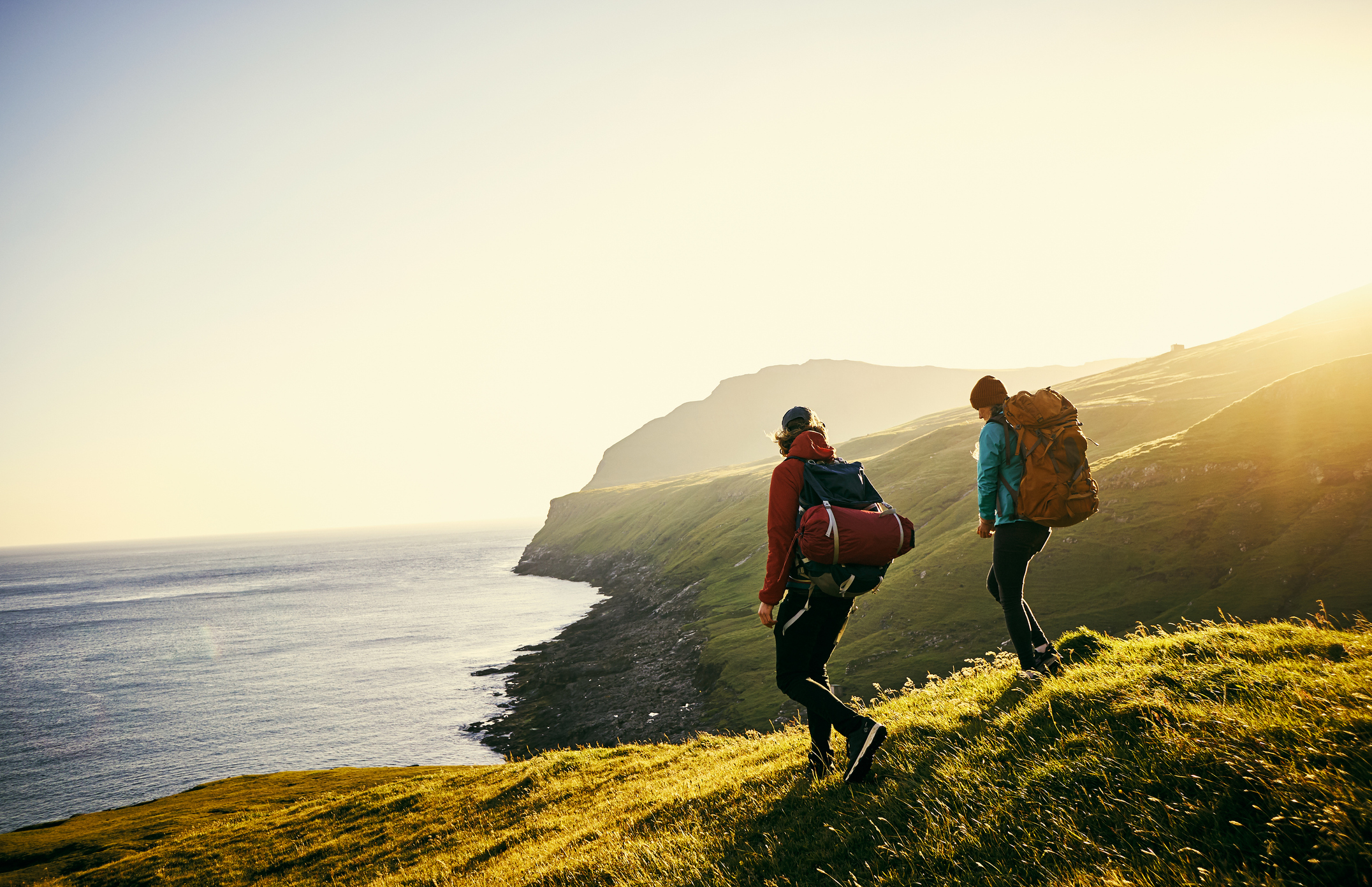 Two hikers with backpacks walk along a grassy hillside, overlooking a calm sea under a bright sunset.