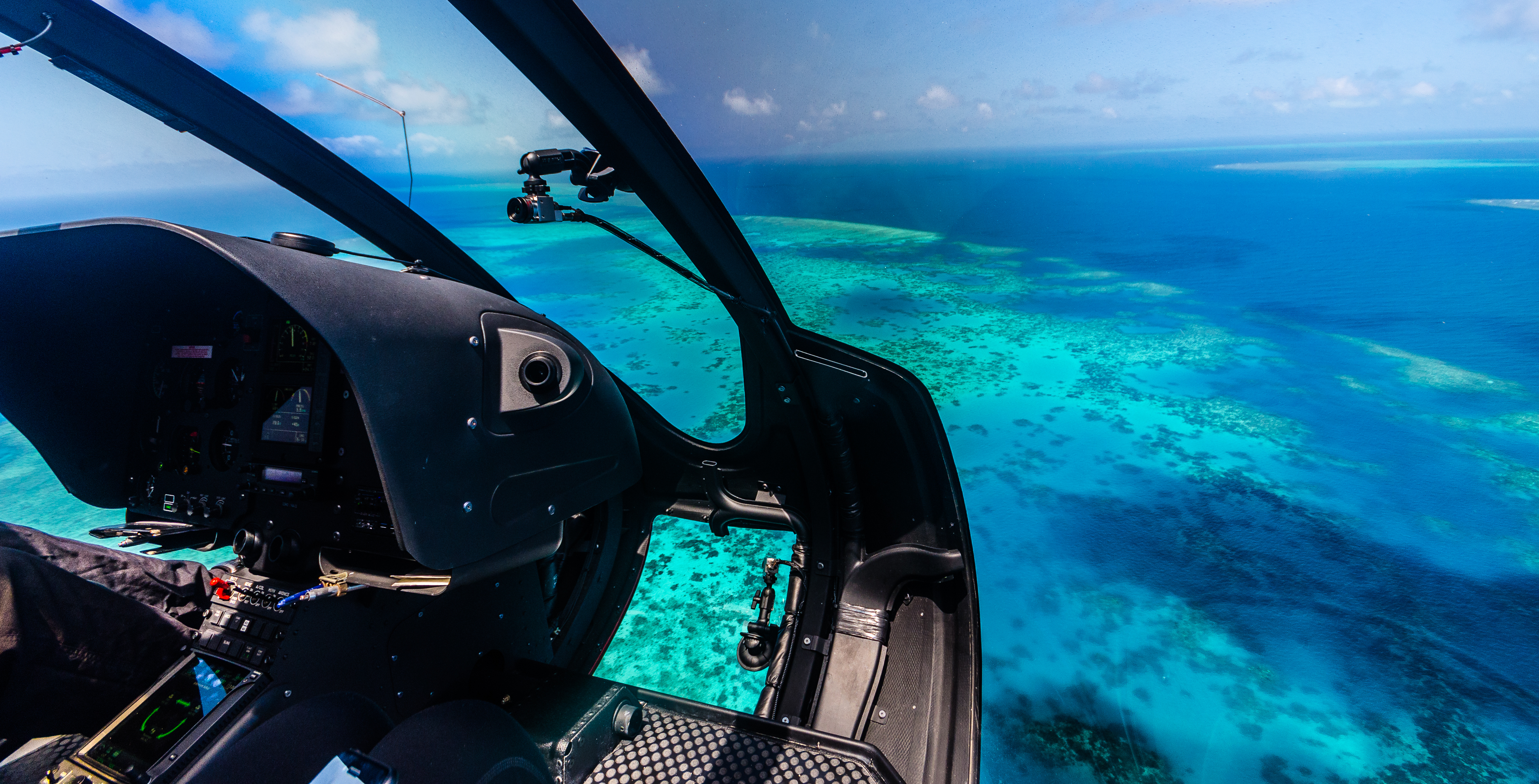 Cockpit view of a helicopter flying over vibrant turquoise waters and coral reefs.