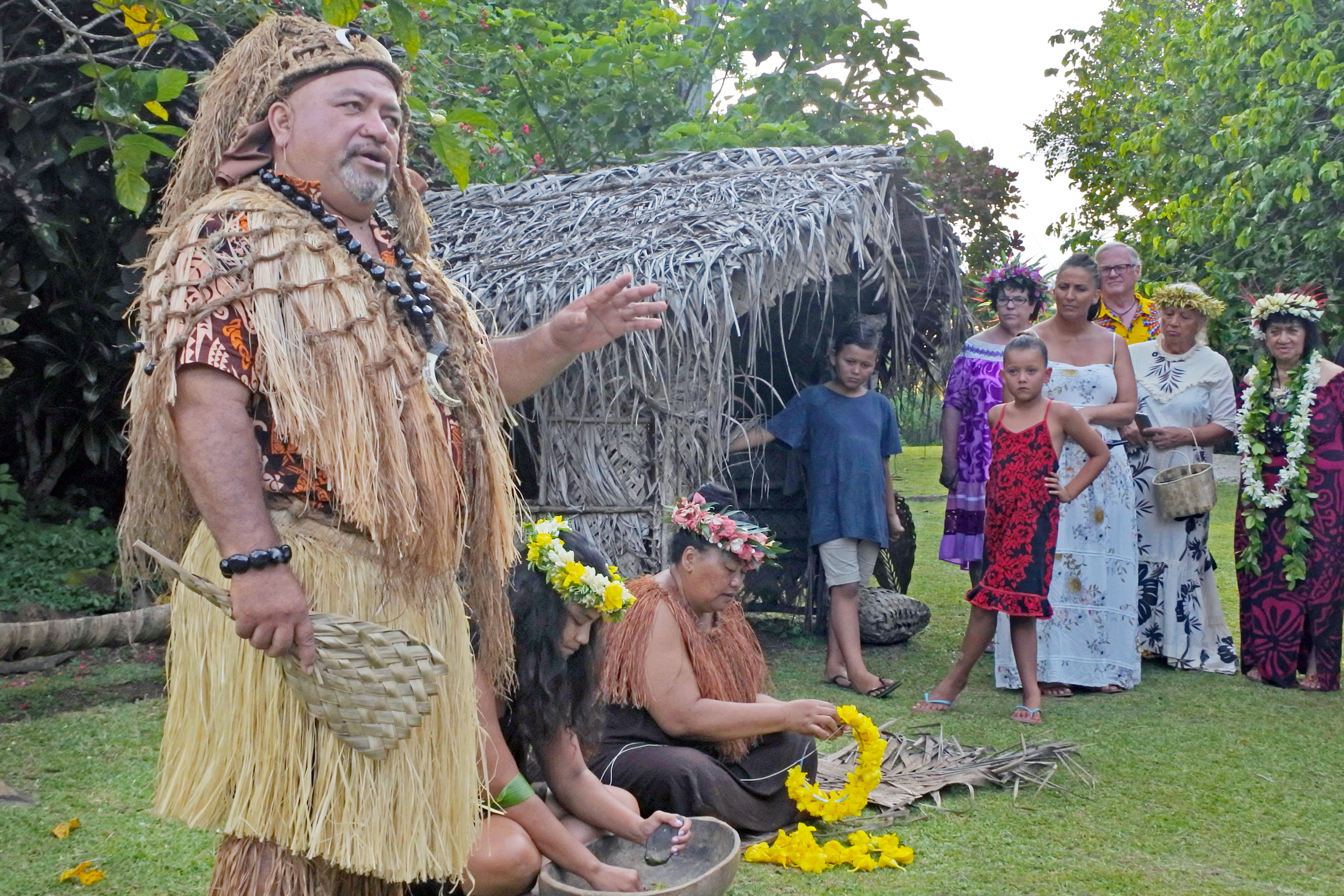 A group of people, including a man in traditional attire, gather near a thatched hut for a cultural presentation.