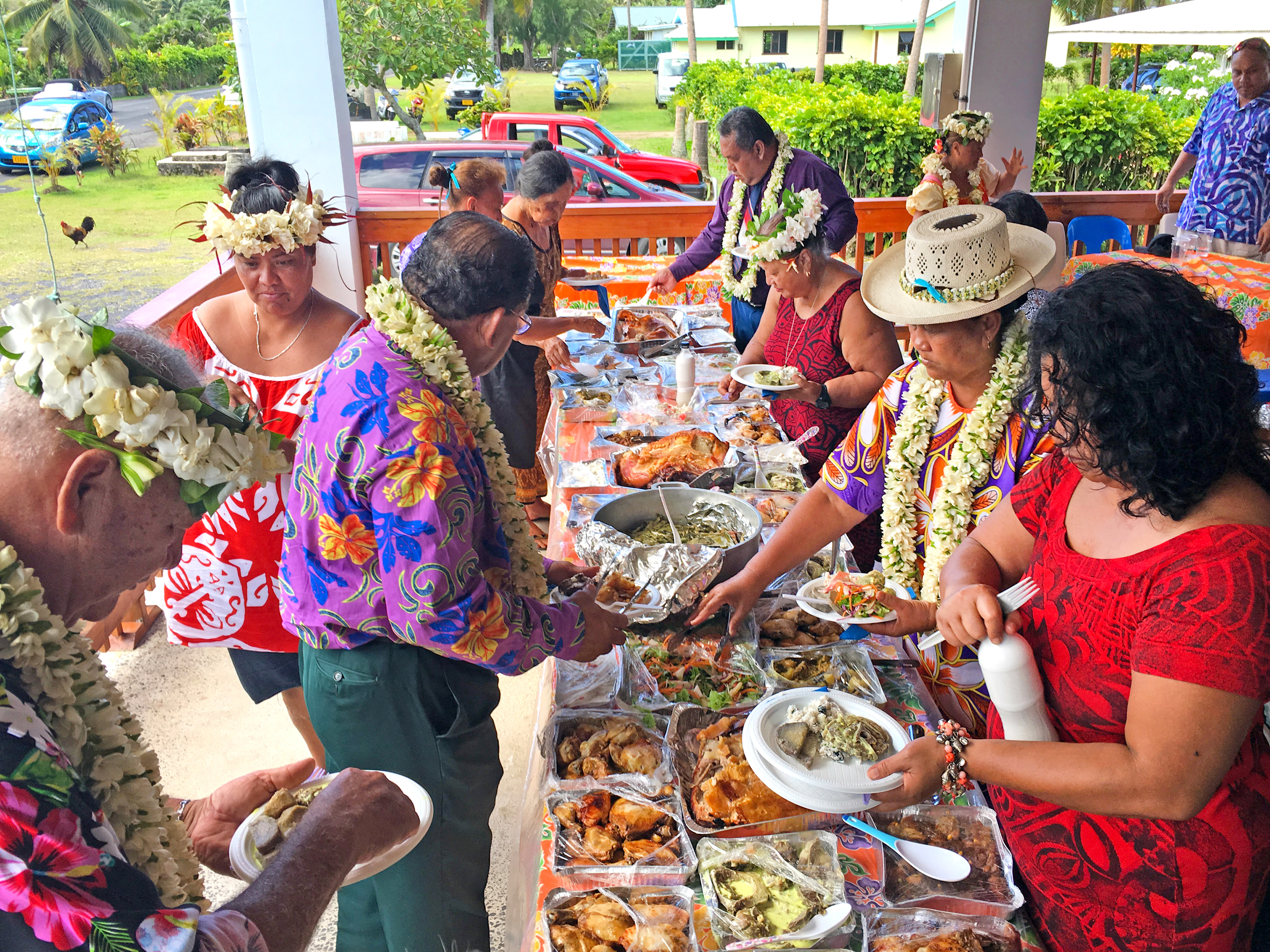 A group of people in colorful attire share a buffet spread outdoors, adorned with leis, enjoying a festive meal together.
